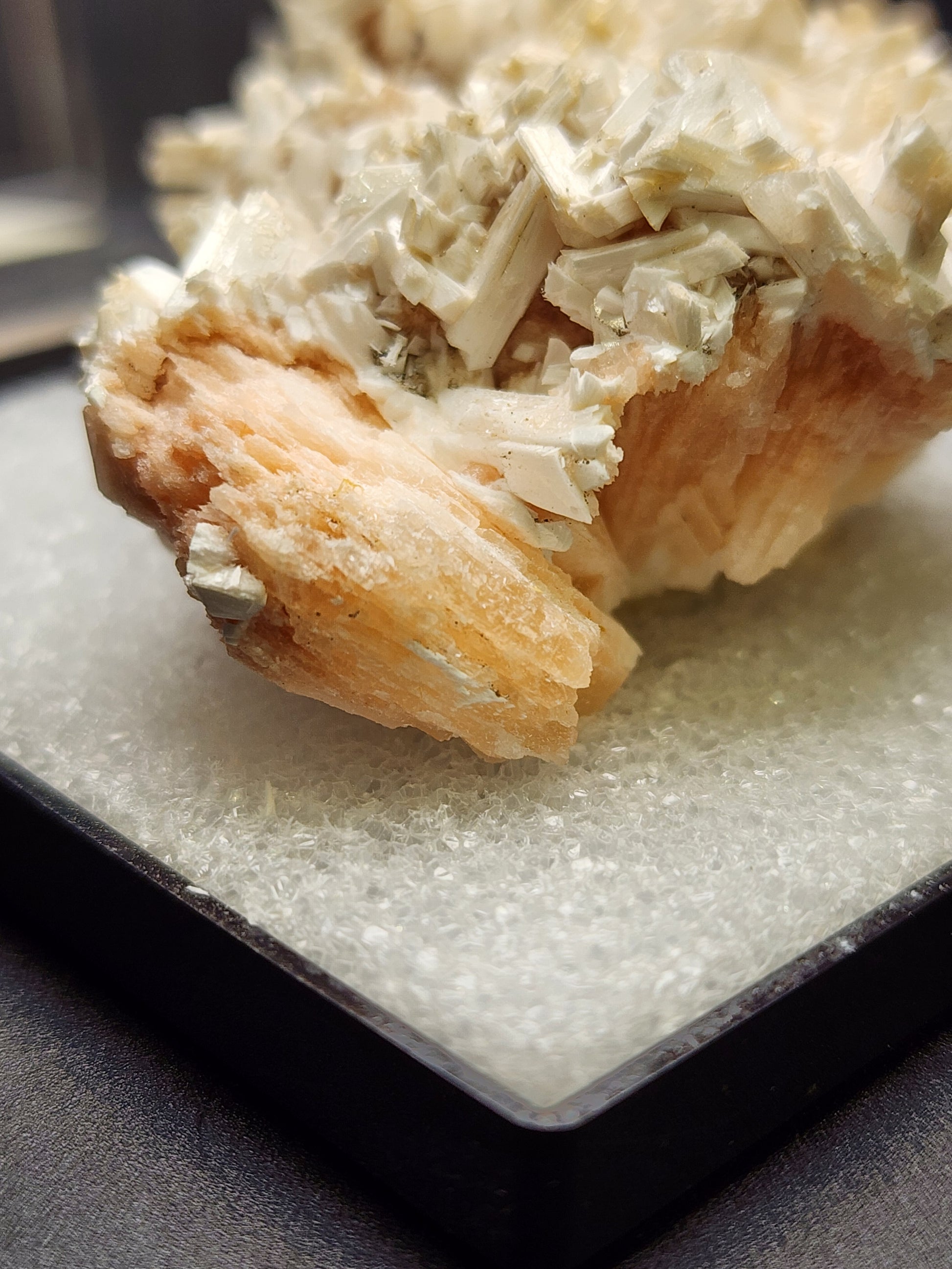 A close-up photograph showcases the AUCTION- Stilbite and Laumontite from Poona, India by The Crystalary. The mineral specimen features pinkish-orange and white crystalline structures resting on a white speckled display surface, highlighting its detailed formations. The blurred background emphasizes the intricate beauty of this exquisite piece.