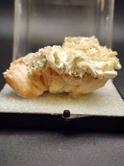 A specimen of Stilbite and Laumontite from Poona, India, predominantly white with hints of peach and beige, is displayed on a flat base covered in fine white granules. The background is blurred to emphasize the detailed texture of the mineral. This stunning piece is available through The Crystalary under the product name AUCTION- Stilbite and Laumontite- Poona, India.