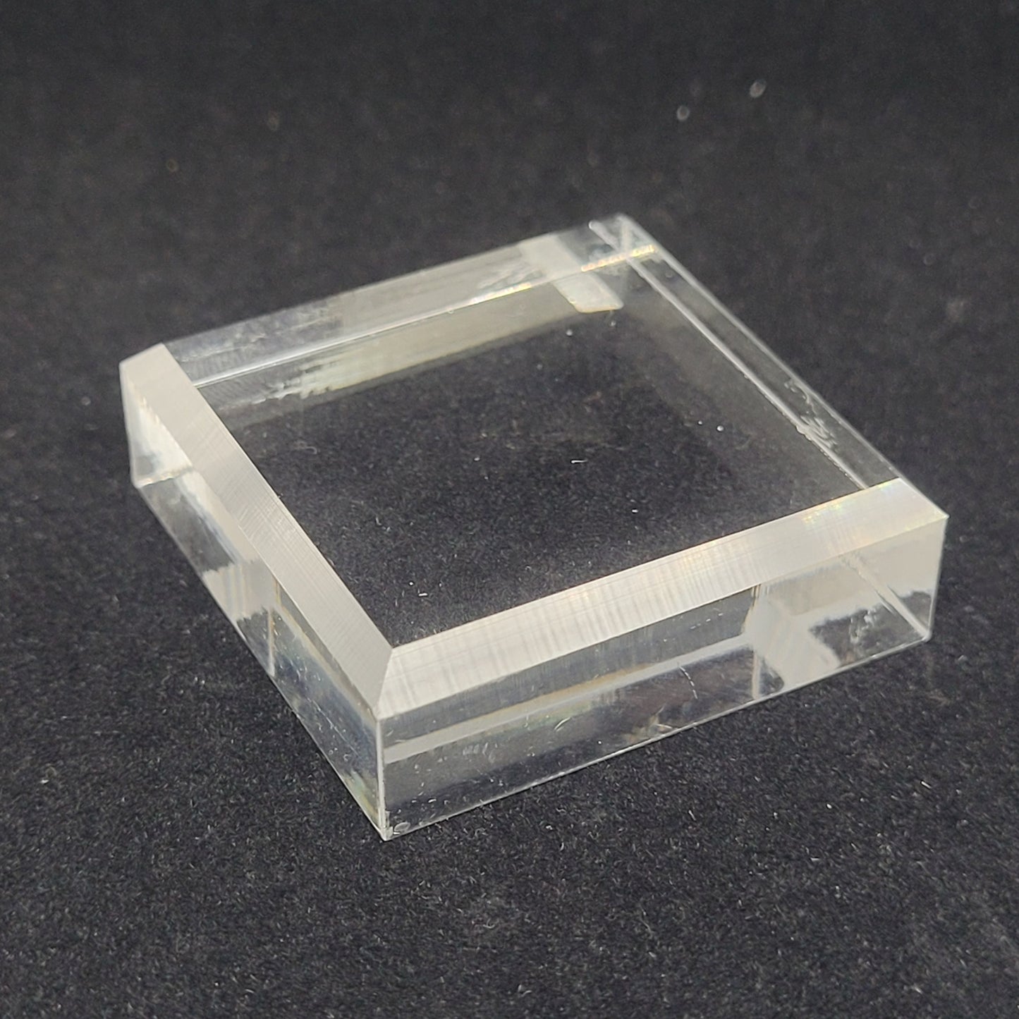 A Beveled Square Acrylic Stand/Mount 1.5" x 1/2" from The Crystalary is placed against a dark background. The flat top surface of the stand allows light to pass through its clear, transparent material, while the slight reflection on its edges adds to its ideal properties for small cabinet mineral displays.