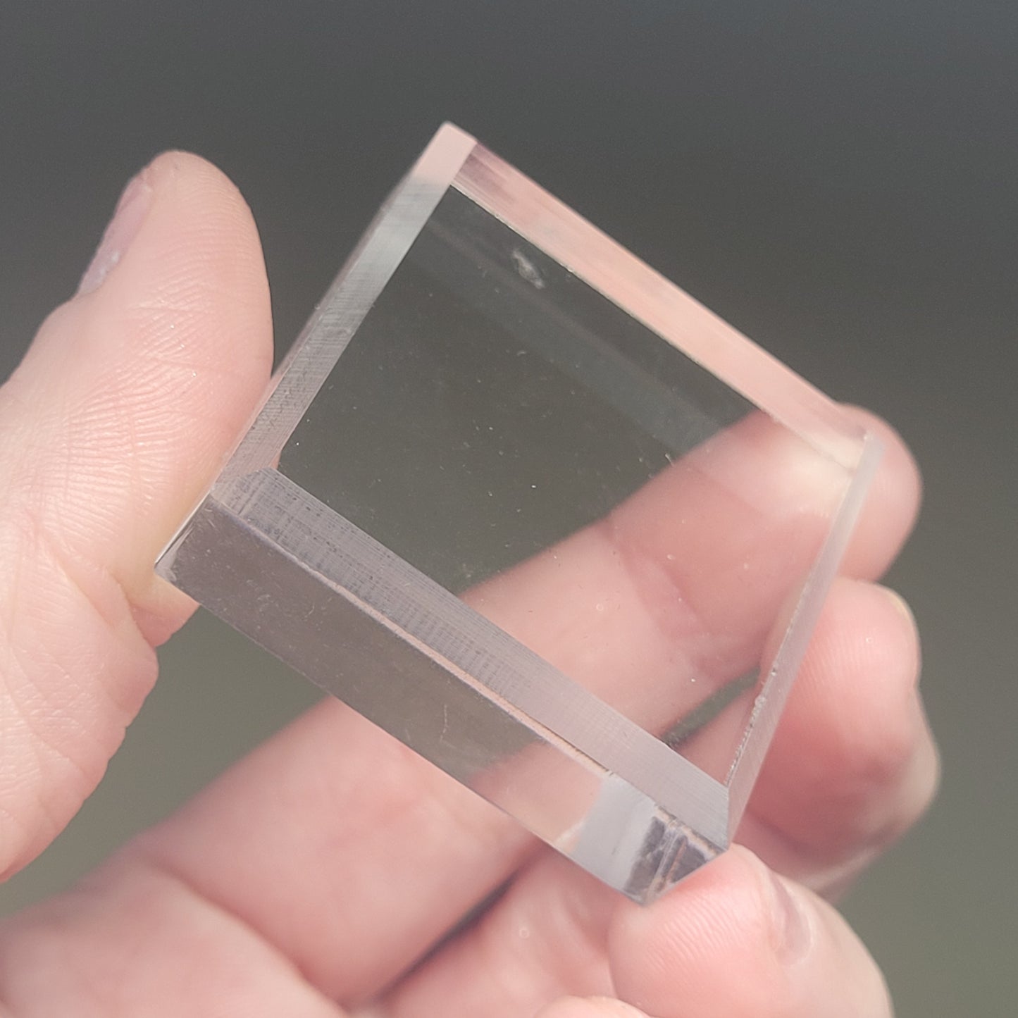 A hand holding a Beveled Square Acrylic Stand/Mount 1.5" x 1/2" from The Crystalary is showcased, much like those used for small cabinet mineral displays. The stand/mount features smooth, polished edges and appears to be made of a clear material like acrylic. The background is blurred.