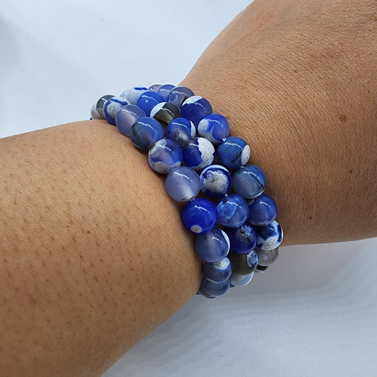 A wrist adorned with The Crystalary's Blue Agate Crystal Bracelet, featuring several strands of blue agate and white marbled beads, forms a stunning accessory. The round, smooth beads are arranged closely together, creating a layered and colorful bracelet that enhances both expressiveness and social skills.