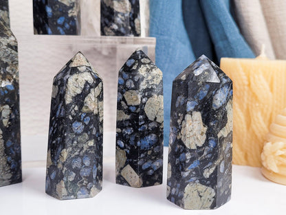 Three polished Blue Que Sera Llanite Points from The Crystalary, showcasing an intricate mix of black, blue, and beige hues, stand elegantly on a white surface. In front of a small acrylic shelf, they are surrounded by candles and fabric in the background. Each stone boasts a unique marbled appearance with varying patterns, chosen intuitively.