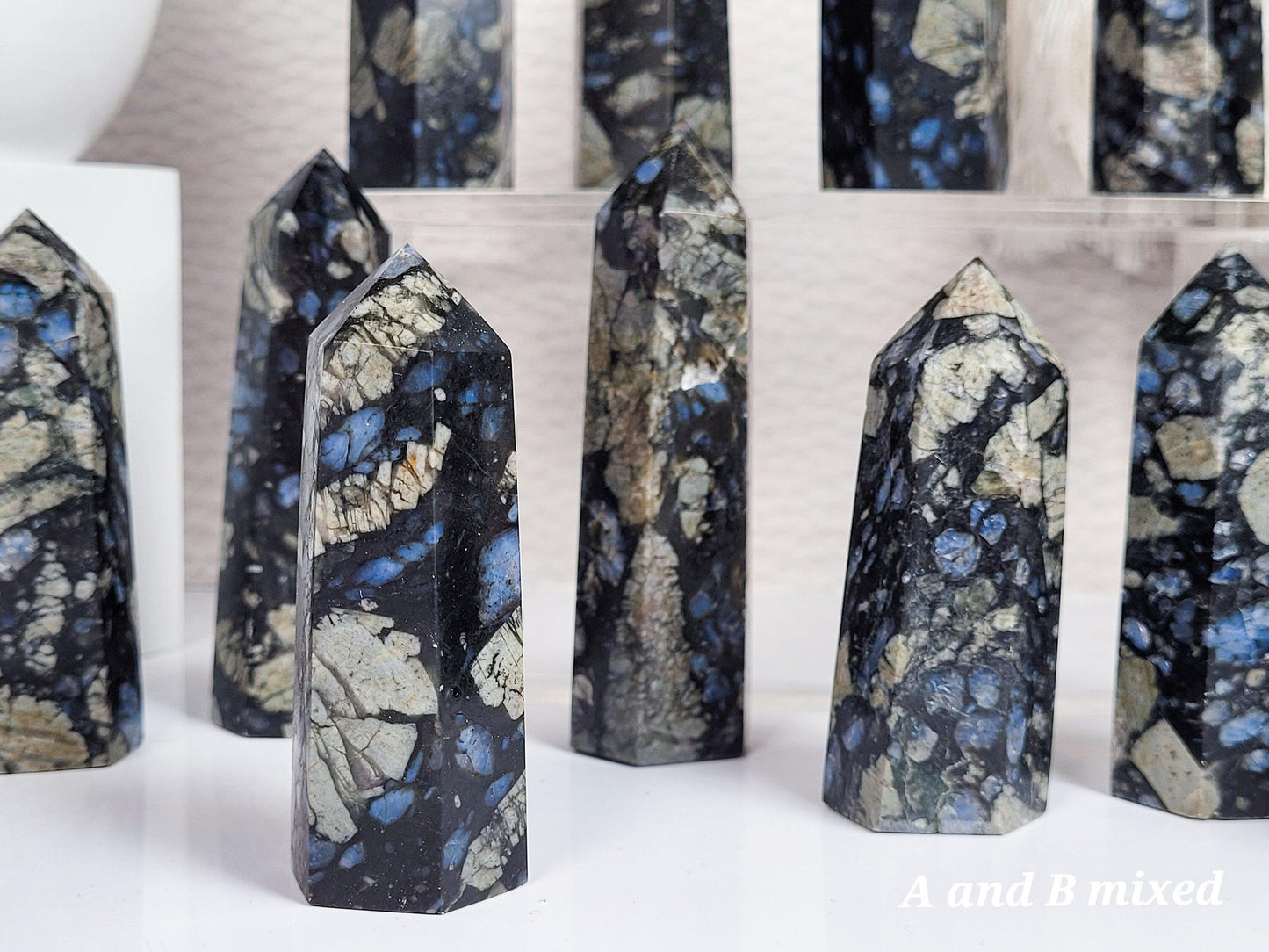 Polished Blue Que Sera Llanite Points from The Crystalary, featuring varying heights and shapes, are displayed. These crystals showcase a mixed pattern of dark earthy tones accented with blue mineral inclusions. Set indoors on a reflective surface, the display includes text reading "A and B mixed" in the corner.