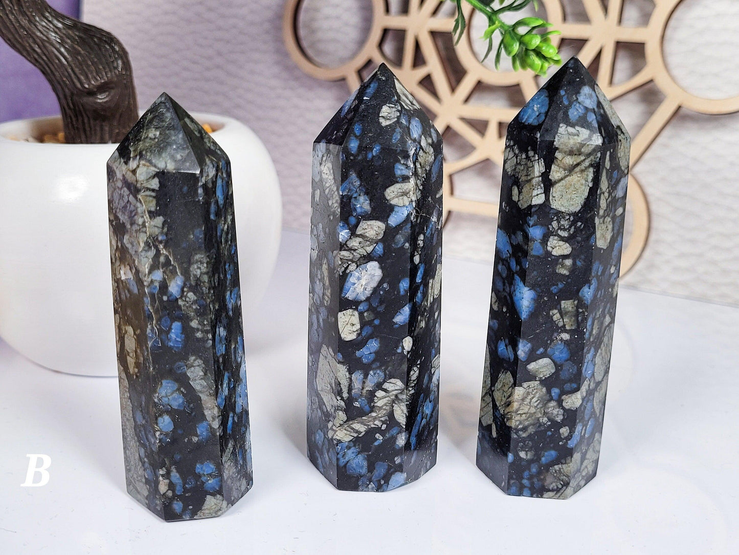 Three hexagonal Blue Que Sera Llanite Points from The Crystalary stand on a white surface. The crystals are dark with flashes of blue, akin to labradorite, and reflective light spots. A white pot with a plant and a circular wooden decoration are in the blurred background. A 'B' is at the lower left corner.