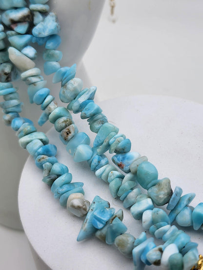 A close-up of The Crystalary's "Blue Sky Quartz & Larimar Chip Bracelet," showcasing irregularly shaped stones in light blue and white hues. The beads feature various shades of blue and white with natural patterns, including quartz inclusions. The bracelet is draped over a white surface.