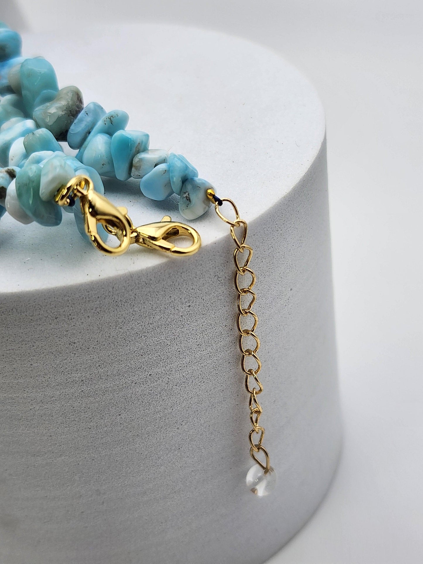 Close-up of the Blue Sky Quartz & Larimar Chip Bracelet by The Crystalary, showcasing a series of irregularly shaped blue-green Larimar gemstones. The clasp and extender chain are gold, ending in a small translucent Quartz bead. The jewelry is displayed against a white, cylindrical background.