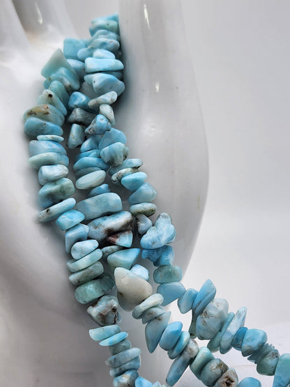 Close-up of smooth, sky-blue Blue Sky Quartz and Larimar gemstone beads strung together and draped over a smooth, white sculpted structure. The stones vary in size and shape, exhibiting a range of light blue tones with occasional white and brown inclusions, reminiscent of The Crystalary's adjustable chip bracelet.