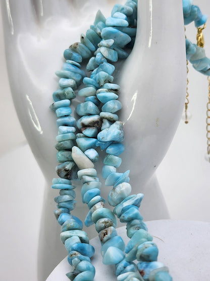 A close-up of a white mannequin bust adorned with multiple necklaces made of polished blue and white gemstone chips, highly reminiscent of turquoise and larimar. The stones vary in size and shape, creating a textured, organic look. An adjustable Blue Sky Quartz & Larimar Chip Bracelet by The Crystalary complements the set. The background is white, highlighting the vibrant colors.