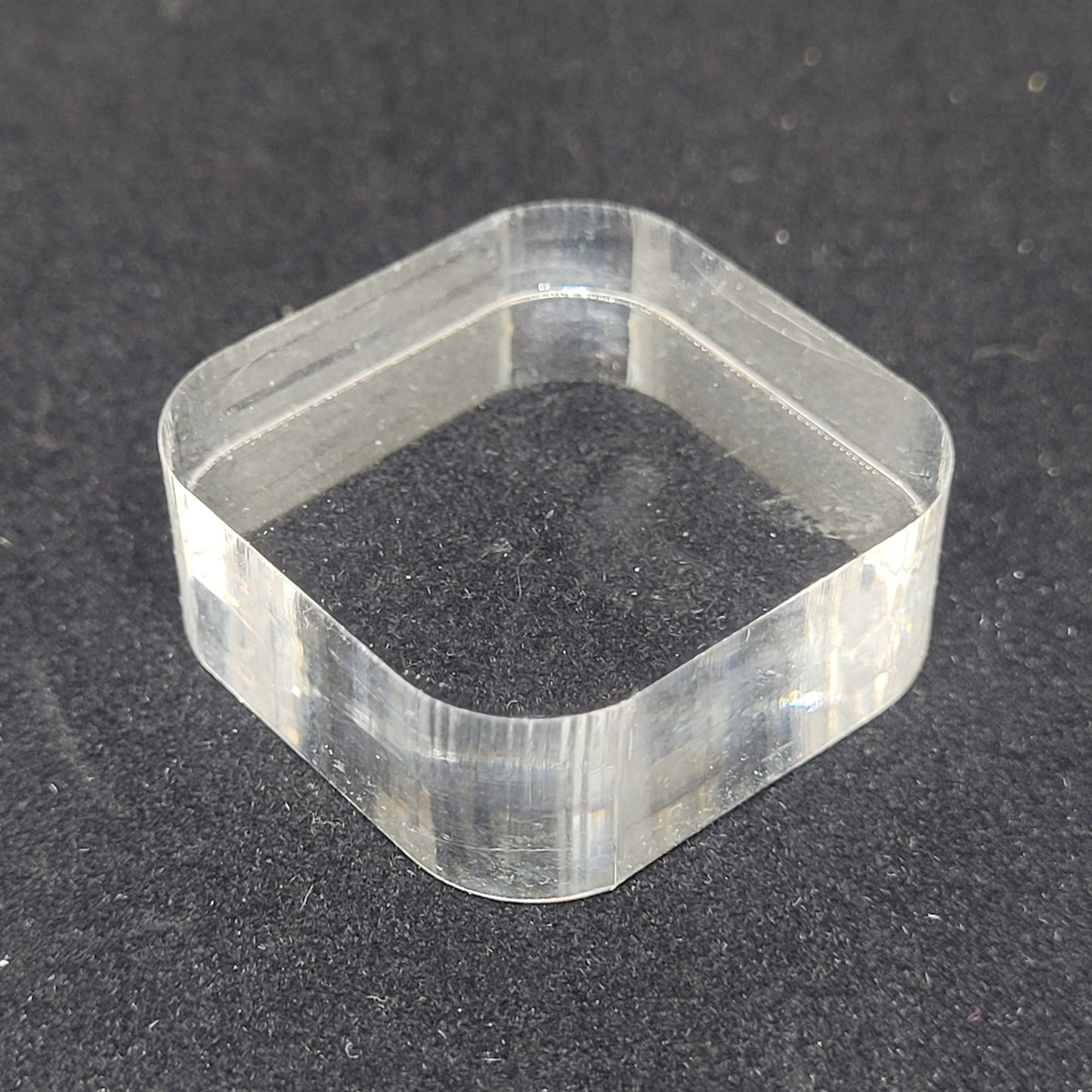 A transparent Bullnose Square Acrylic Stand/Mount from The Crystalary, measuring 1 3/16" x 1/2", lies flat on a black surface. The stand/mount has rounded corners and smooth edges, perfect for use with Mini Mineral displays.