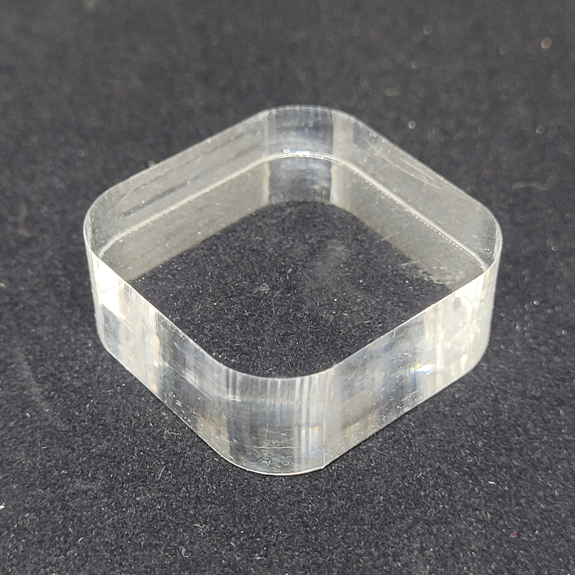 A transparent Bullnose Square Acrylic Stand/Mount from The Crystalary, measuring 1 3/16" x 1/2", lies flat on a black surface. The stand/mount has rounded corners and smooth edges, perfect for use with Mini Mineral displays.