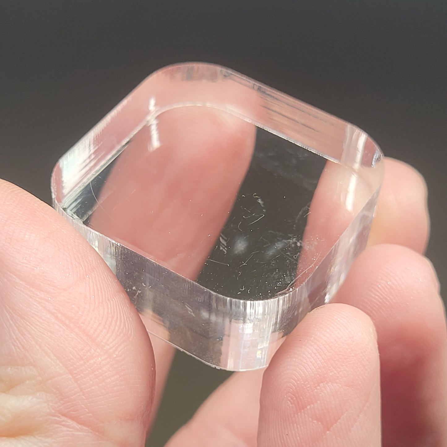 Close-up of a hand holding a small, transparent, rectangular piece identified as the Bullnose Square Acrylic Stand/Mount 1 3/16" x 1/2" by The Crystalary. The material appears smooth with rounded edges, reflecting light and showing minor surface scratches. Ideal for mini mineral displays. The background is blurred and out of focus.