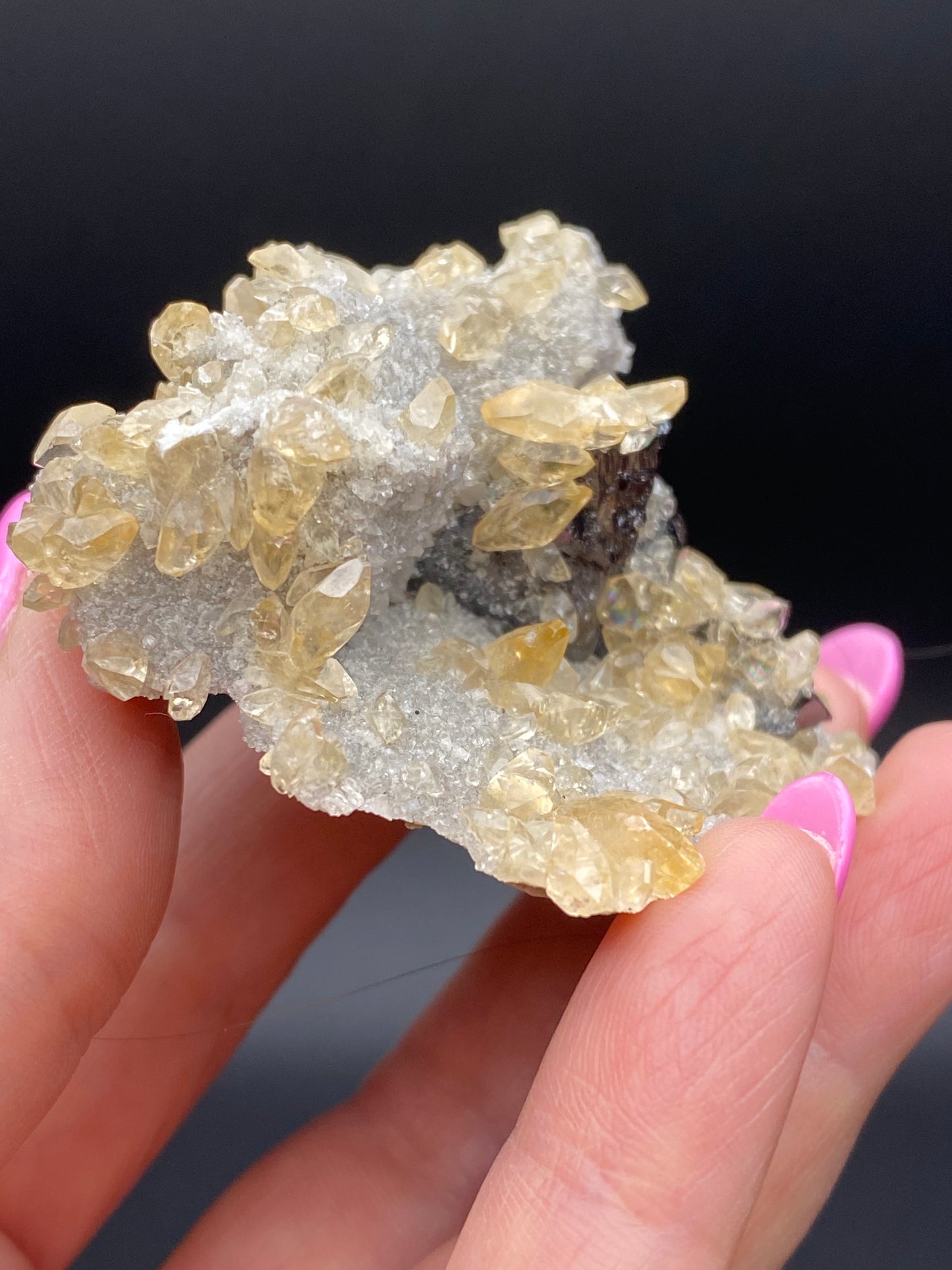 A close-up shot captures a hand holding a cluster from The Crystalary's Calcite, Quartz, and Sphalerite collection, sourced from the Elmwood Mine in Carthage, Smith County, Tennessee. The striking light yellow crystals stand out against their rough gray base. Pink painted nails provide a vivid contrast to the clear, slightly yellow-tinted formations. A dark background enhances the intricate texture and details of these captivating crystals.