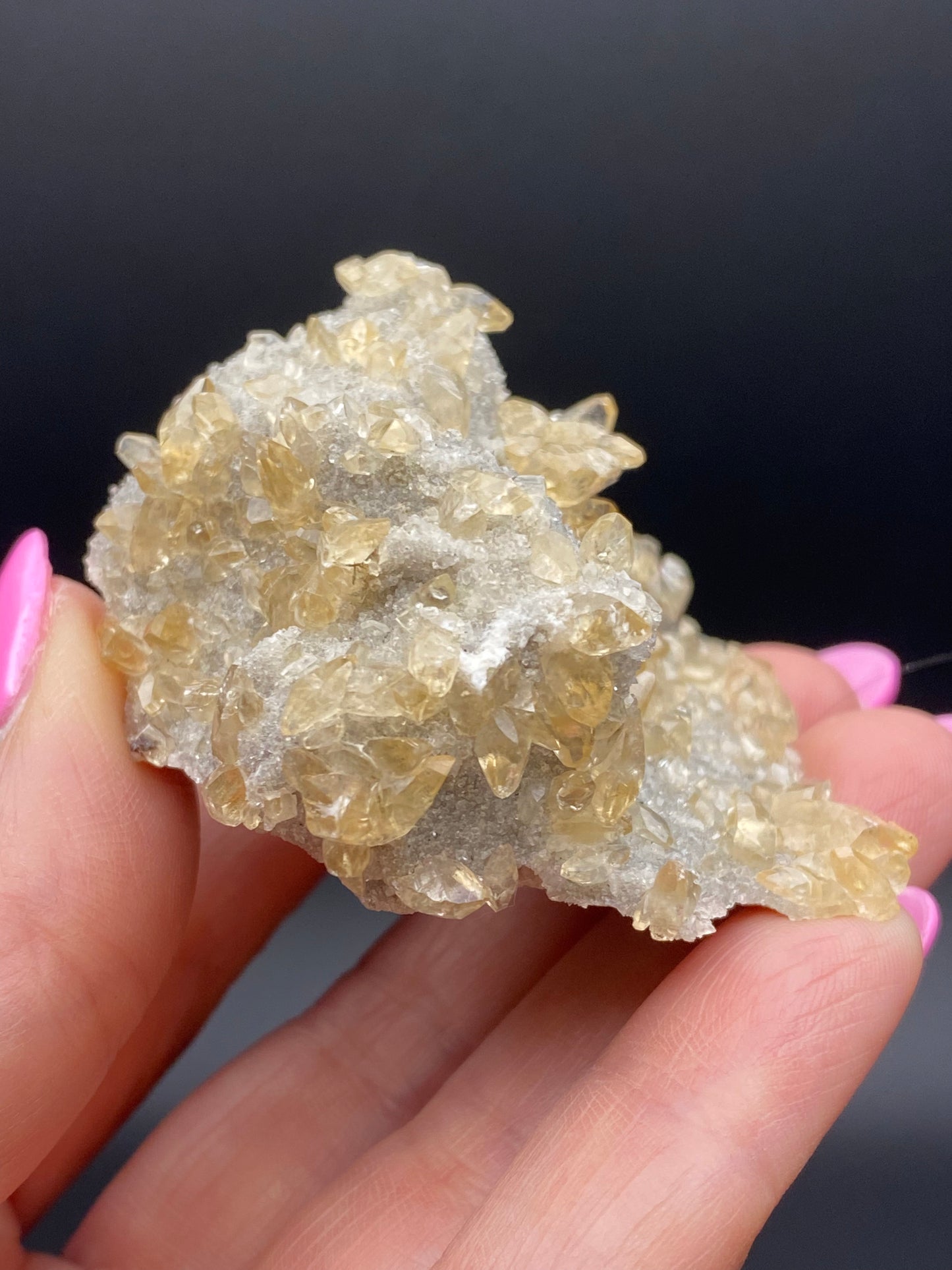 A close-up of a hand holding a cluster of Calcite, Quartz, and Sphalerite from the Elmwood Mine in Carthage, Smith County, Tennessee. Provided by The Crystalary, the crystals exhibit a pale yellow hue with tiny druzy quartz against a dark background. They are small, translucent, and densely packed, creating a rough, textured appearance. The hand's fingernails are painted pink.