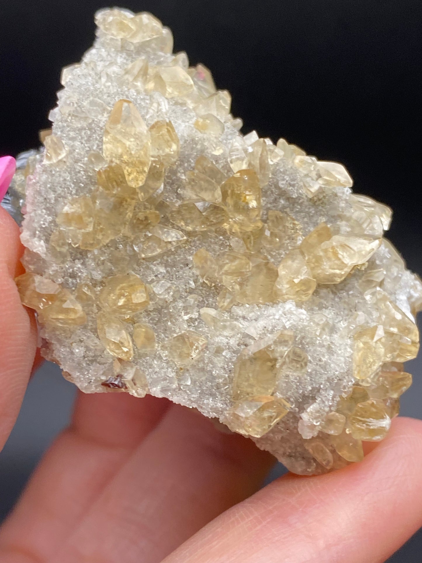 Close-up image of a hand holding a "Calcite, Quartz and Sphalerite" mineral cluster from The Crystalary. The cluster, sourced from Elmwood Mine in Carthage, Smith County, Tennessee, USA, consists of many small, pale yellow translucent crystals embedded in a rough white matrix. Gleaming calcites are visible among them. One hand has visible pink fingernail polish against an out-of-focus and dark background.