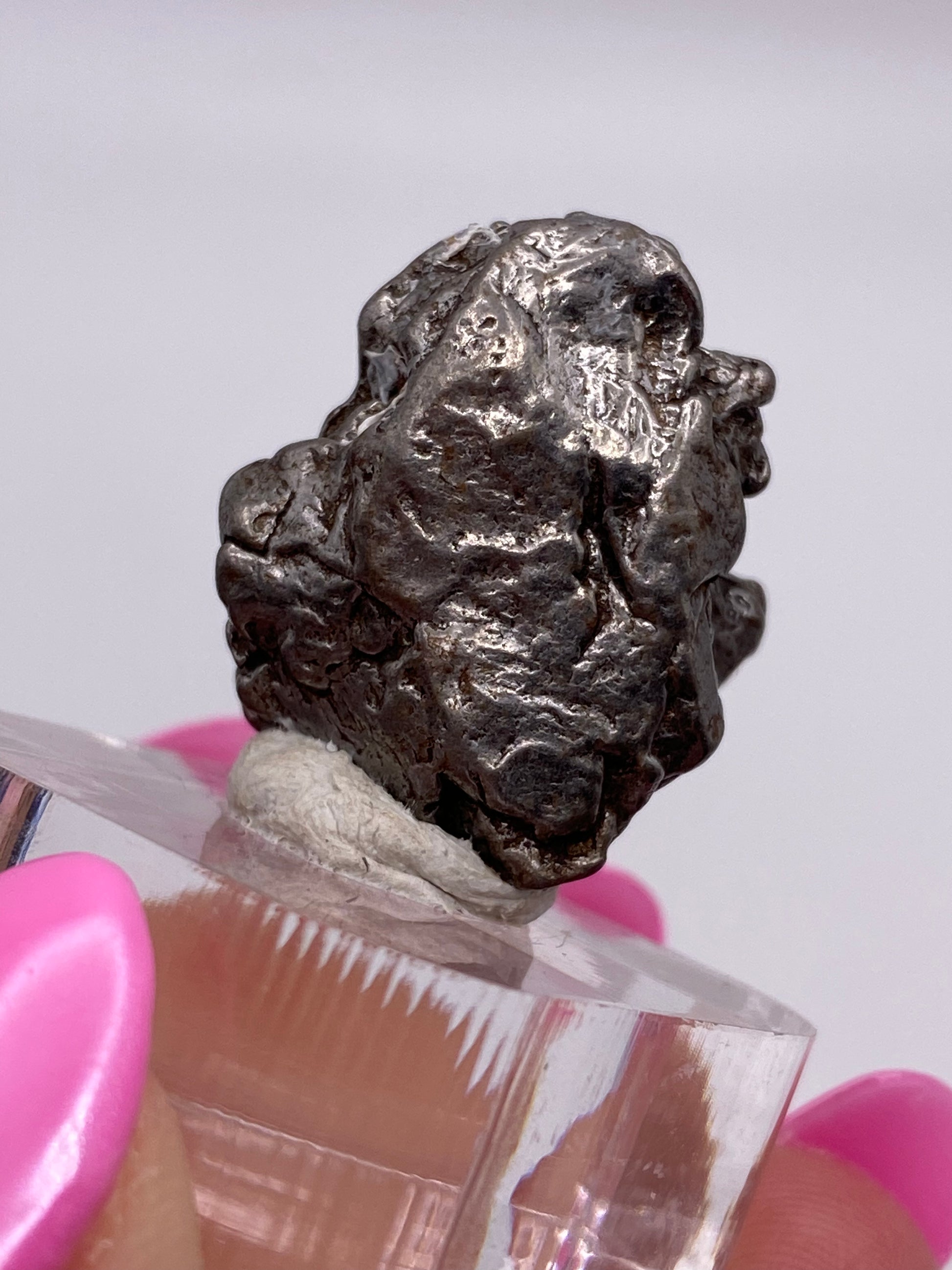 A small metallic nugget, believed to be a Campo del Cielo Meteorite from Gancedo, Doce de Octubre Department, Chaco Province, Argentina, is displayed on a transparent stand. The rough, irregular surface of this iron meteorite from The Crystalary glistens as it's held by a hand with bright pink acrylic nails against a blurred, light-colored background.