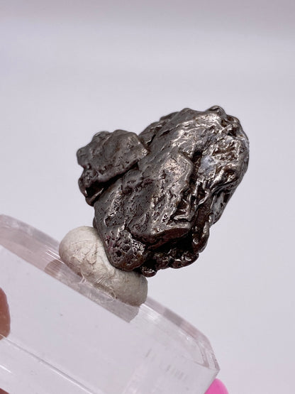 A close-up view of a metallic silver Campo del Cielo Meteorite from The Crystalary, originating from Gancedo in the Doce de Octubre Department, Chaco Province, Argentina. Displayed on a clear plastic stand and supported by a piece of white putty, this meteorite features a rough, textured surface and an uneven shape. The background is blurred and minimalistic.