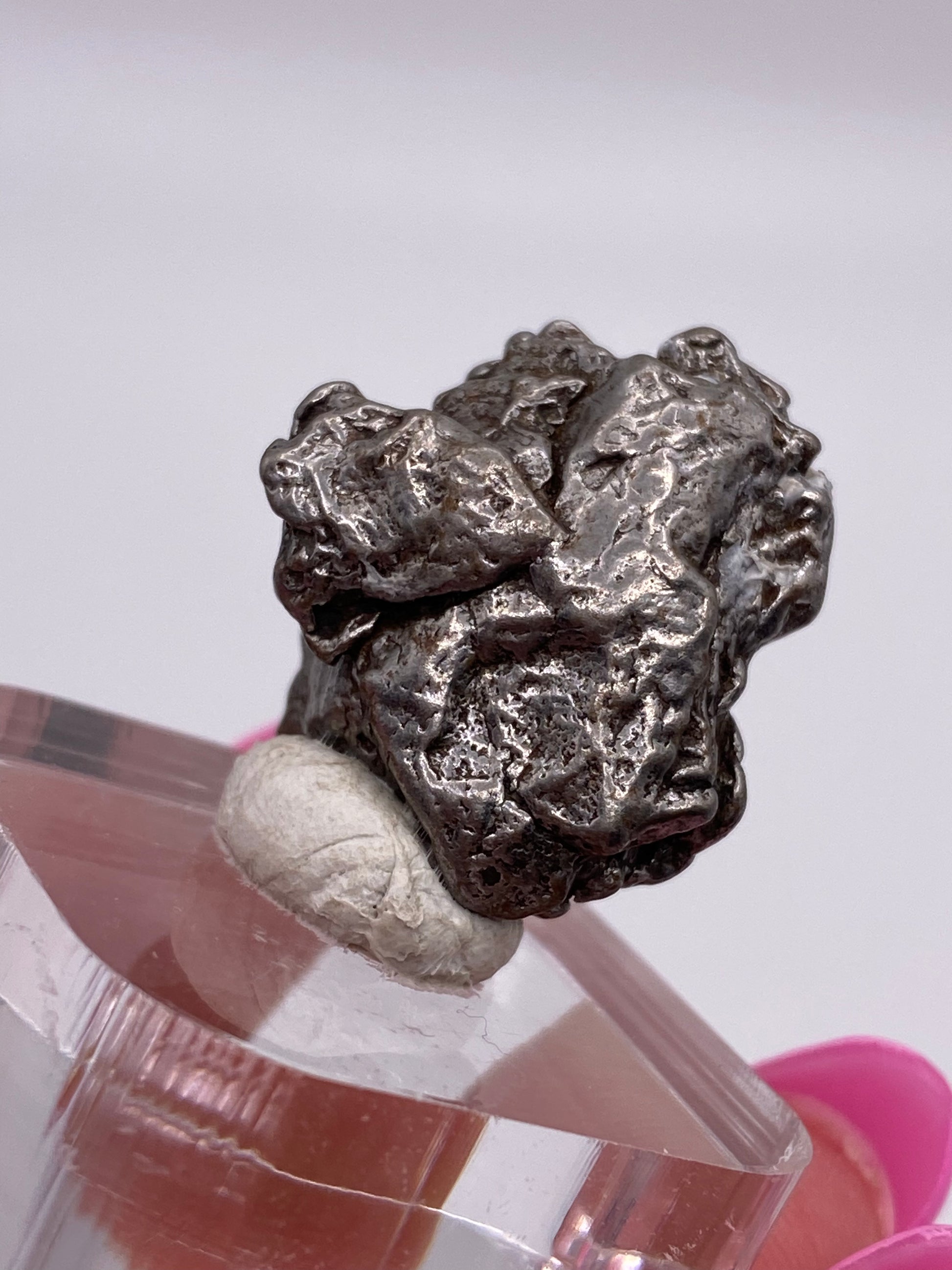 A close-up image shows an irregularly shaped, metallic silver nugget identified as a Campo del Cielo Meteorite from The Crystalary, specifically sourced from Gancedo, Doce de Octubre Department, Chaco Province, Argentina. It is displayed on a clear plastic base. The background is out of focus, and a hand with bright pink nails is visible holding the base.