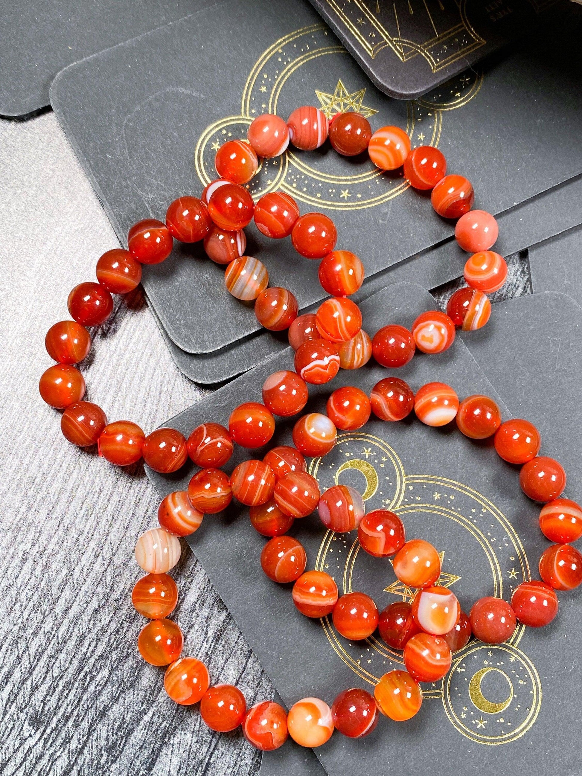 A close-up of three Carnelian Crystal Bracelets from The Crystalary, featuring polished red and orange agate beads, placed on black tarot cards adorned with gold celestial designs. The table has a wood grain texture, showcasing the beads' various stripe and swirl patterns.