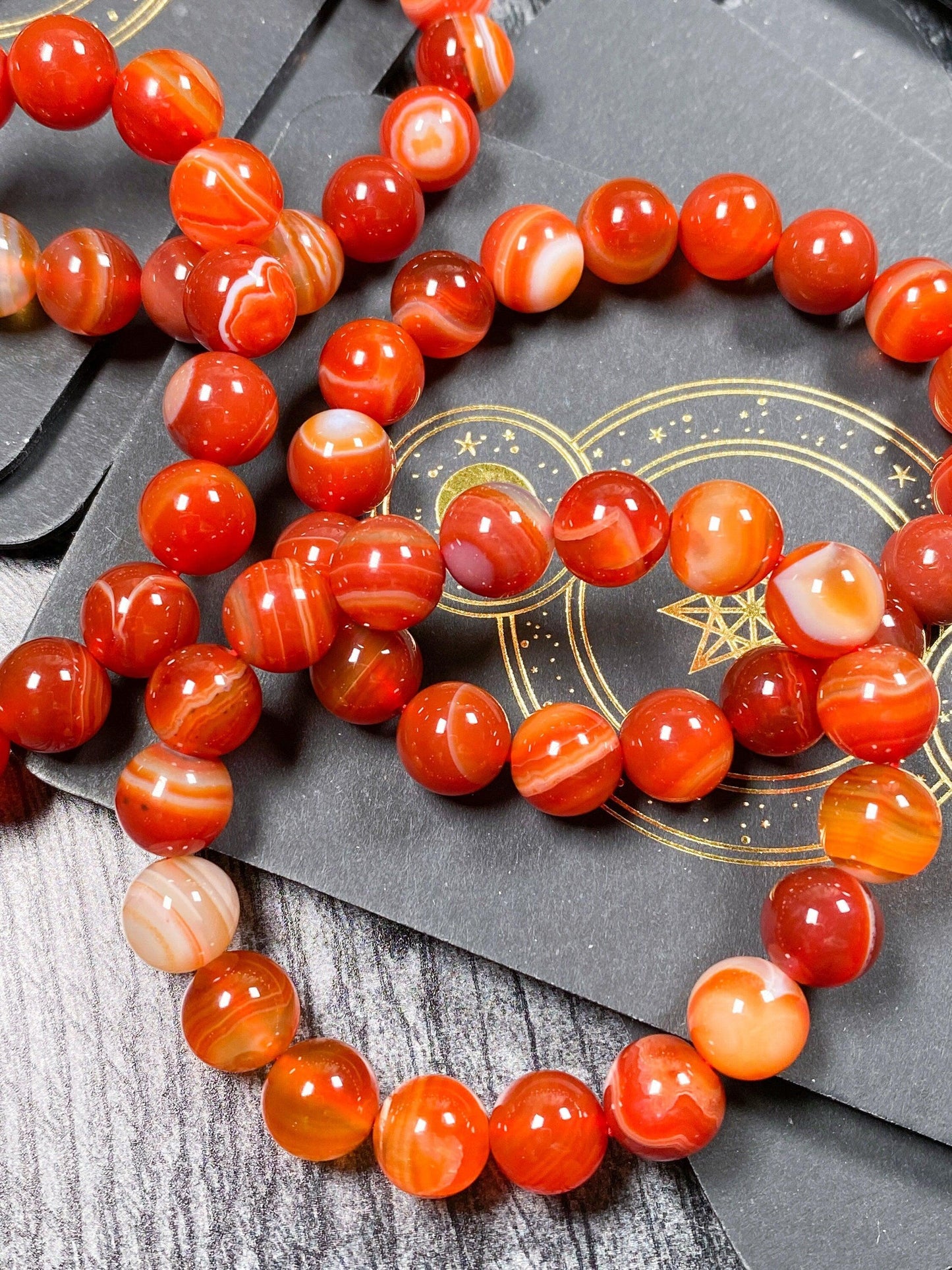 A close-up image showcases a Carnelian Crystal Bracelet by The Crystalary, featuring polished red and orange agate stones with intricate swirling patterns. Resembling fine Carnelian jewelry, the bracelet is displayed on a dark grey board adorned with golden geometric designs. The detailed texture and bead width are clearly visible.