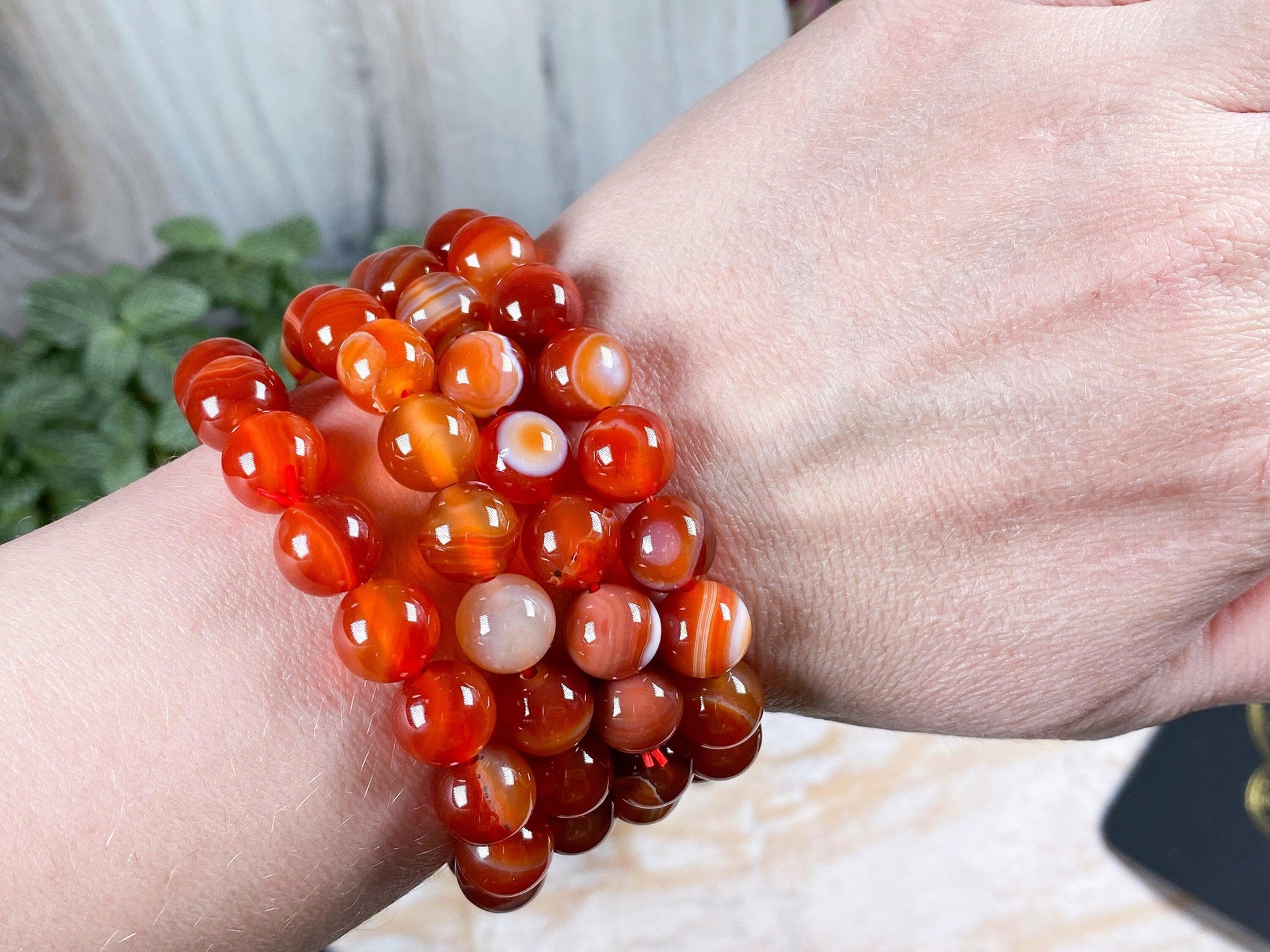 A person's wrist adorned with multiple strands of The Crystalary's Carnelian Crystal Bracelet, showcasing polished, round, orange-red carnelian gems. The background features green foliage and a wooden surface.