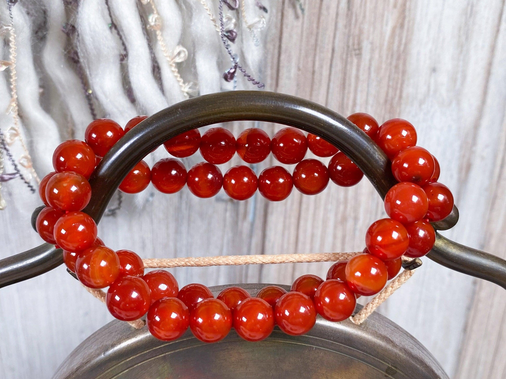 Two Carnelian Crystal Bracelets by The Crystalary, made of round, polished red-brown stones, are draped over a dark metal handle. The background features a rustic wooden surface adorned with textured decorations.