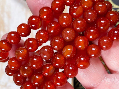 Close-up of a person's hand holding several strands of polished, round beads from The Crystalary's Carnelian Crystal Bracelet. The beads have a glossy finish with some variation in their coloration, showcasing deep reds with hints of orange. This exquisite piece is an example of fine Carnelian jewelry.