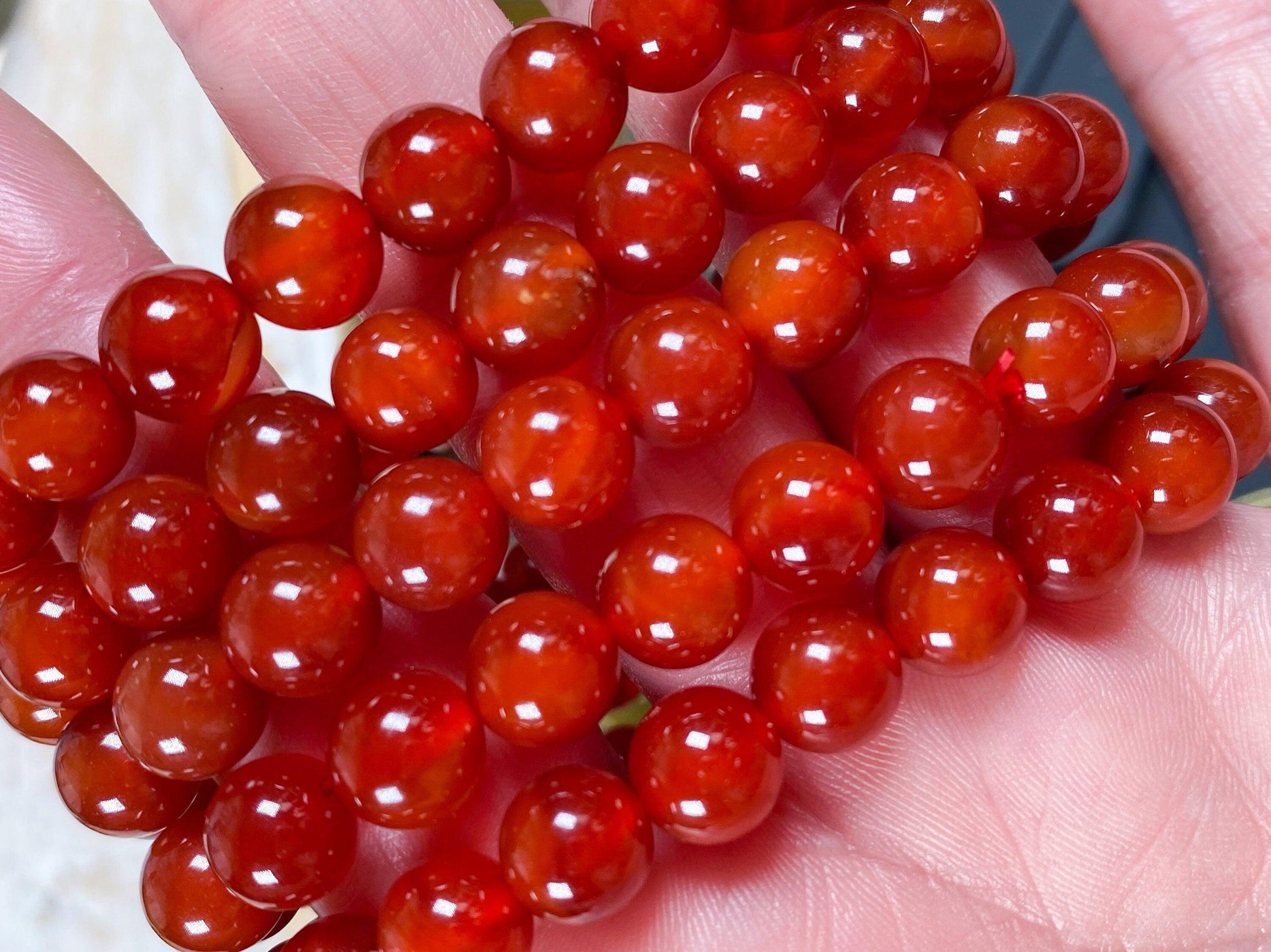 A hand holds a string of polished red beads, each bead appearing smooth and glossy. The beads have a rich, deep red color and are closely packed together, showcasing their shiny surface and uniform round shape. This exquisite Carnelian Crystal Bracelet by The Crystalary highlights the beauty of natural crystal jewelry.