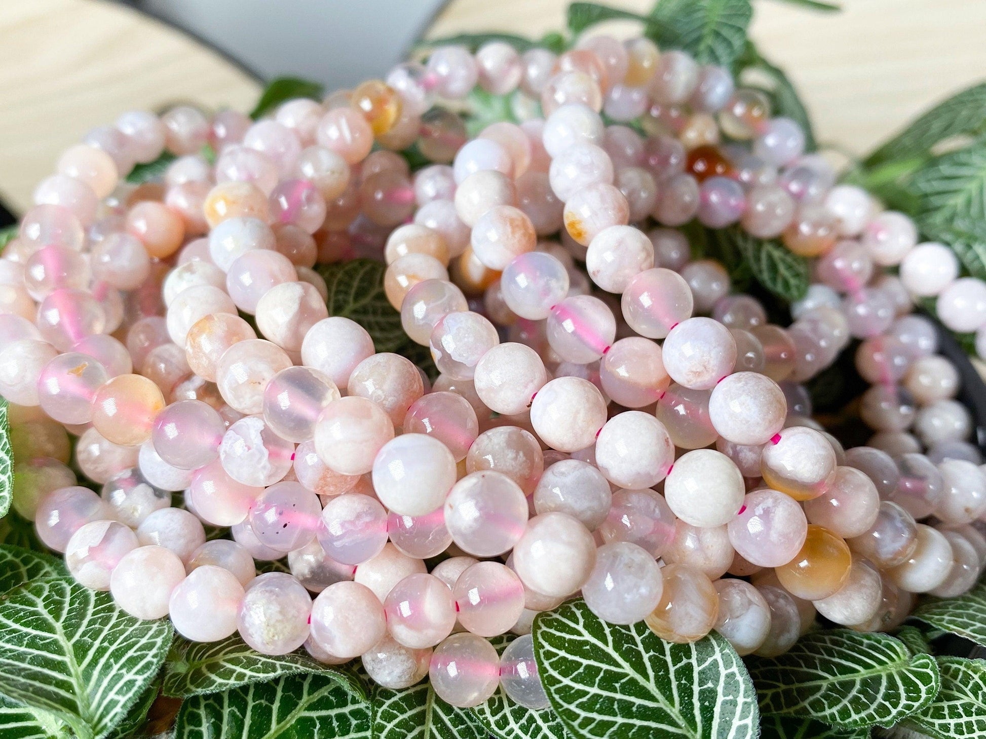 The Crystalary's Cherry Blossom Flower Agate Crystal Bracelets, arranged in several coils, are displayed on a bed of green leaves with a white vein pattern. The glossy beads exhibit varying shades of pastel pink and pale white, reminiscent of delicate cherry blossoms.