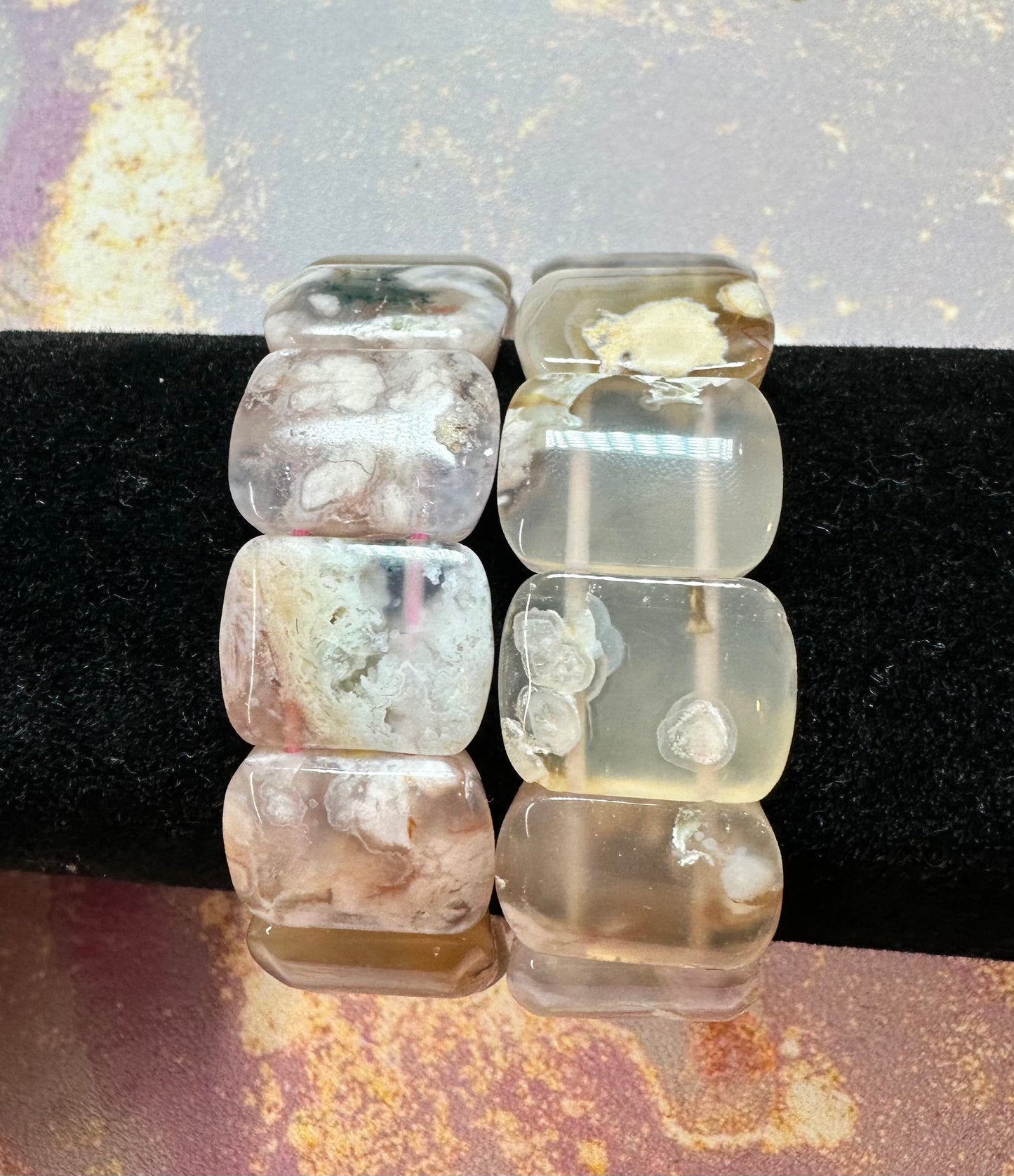 Two crystal bracelets by The Crystalary are displayed on a black, velvety surface. One bracelet, the Cherry Blossom Flower Agate Crystal Bracelet, features large, polished, rectangular beads with cloud-like inclusions. The other bracelet showcases clearer beads with visible natural patterns. The background is mottled with pastel colors.