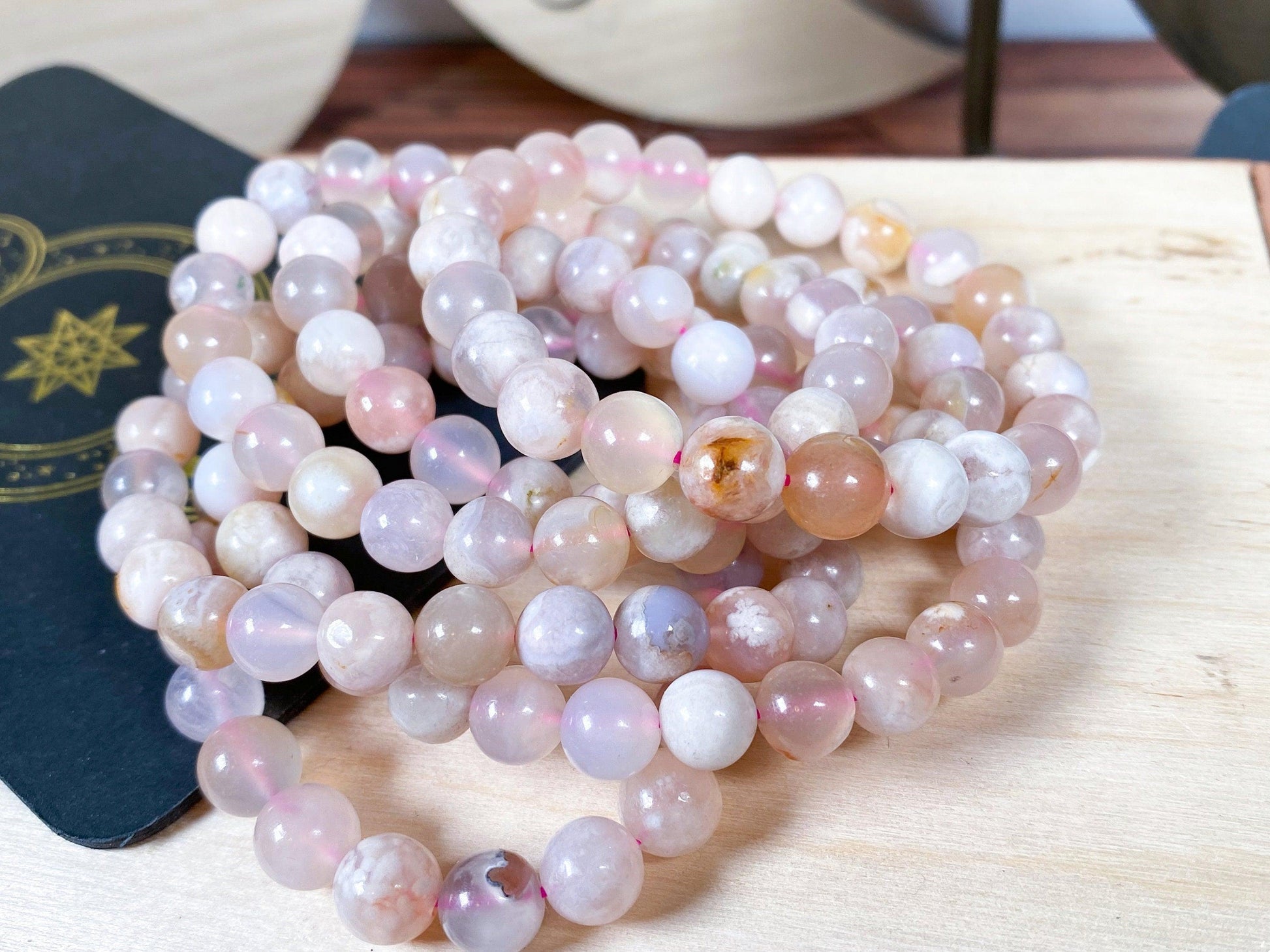 A collection of Cherry Blossom Flower Agate Crystal Bracelets by The Crystalary, featuring polished pink, white, and beige gemstone beads in various shades, intricately strung together. The bracelets rest on a light wooden surface, with a decorative black card showcasing a gold star-like design partially visible in the background.