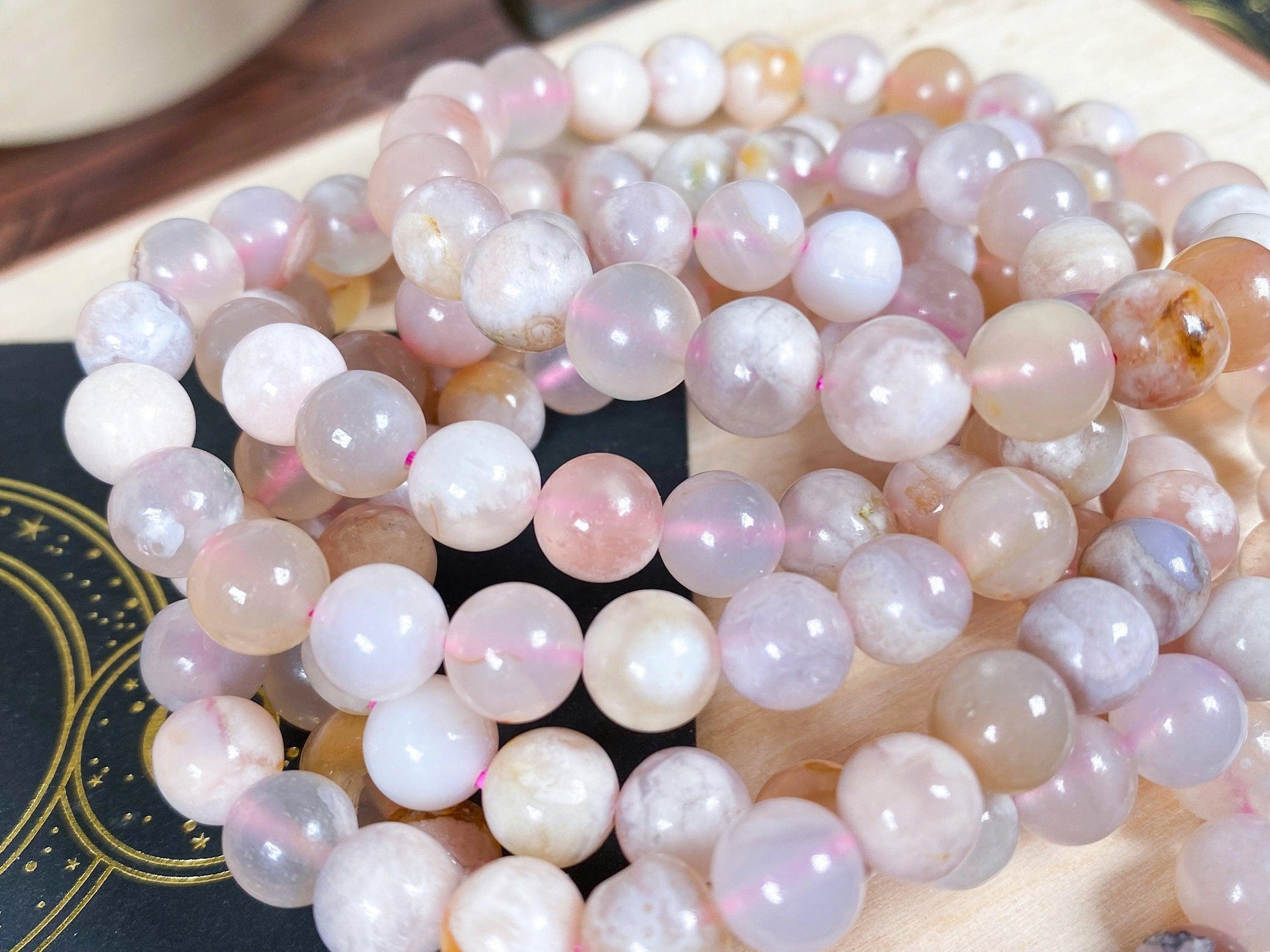 A close-up of several Cherry Blossom Flower Agate Crystal Bracelets from The Crystalary. These bracelets feature translucent, pale pink beads with slight variations in color and pattern, ideal for balancing the heart chakra. They are coiled together and set against a soft beige background with a glimpse of a celestial-themed black object.