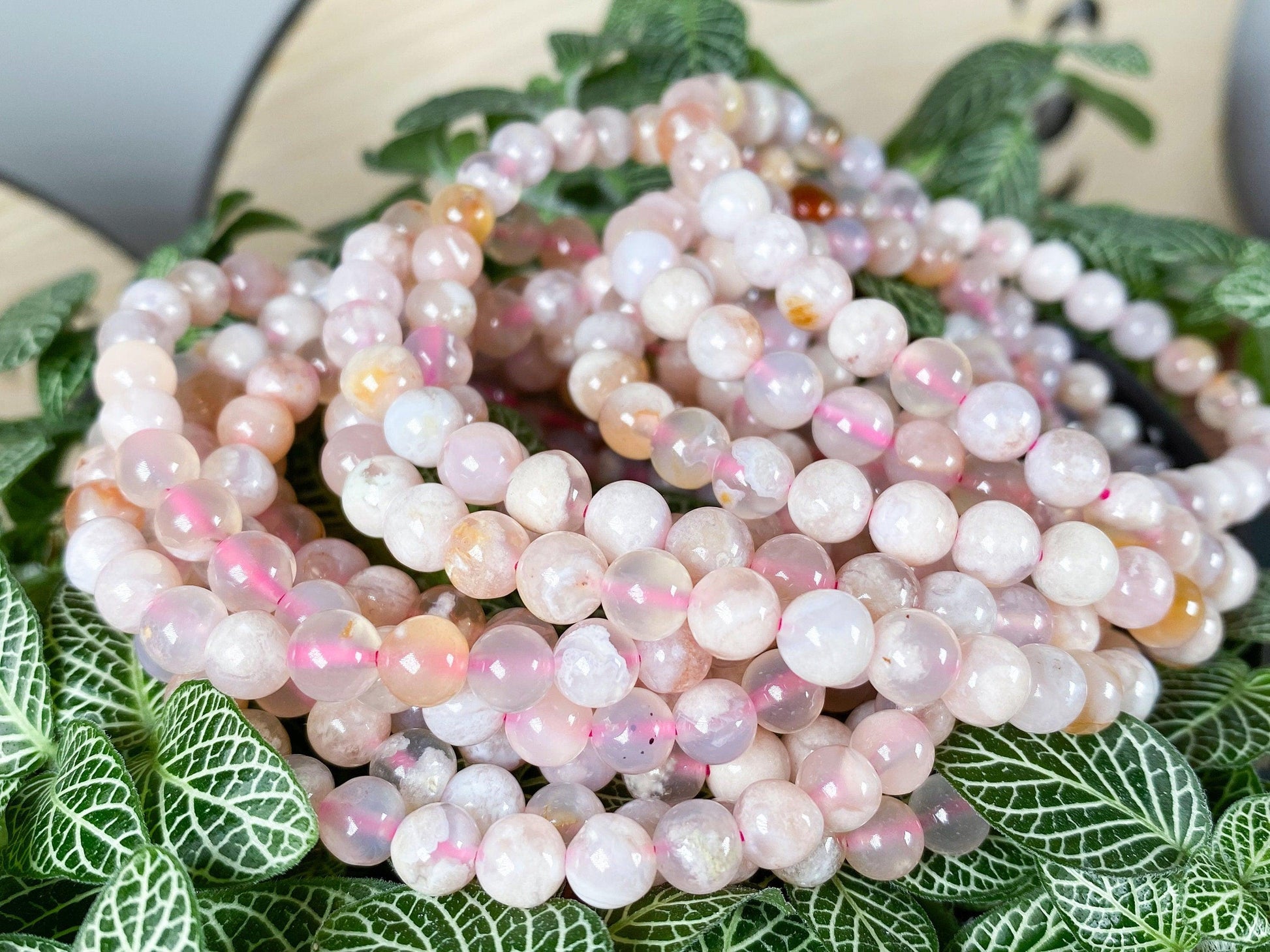 A close-up of multiple strands of round, pale pink beads arranged on top of green, leafy plants. The semi-translucent beads display subtle variations in color, ranging from light pink to white. Like Cherry Blossom Flower Agate Crystal Bracelets by The Crystalary, they exude a soothing beauty against the patterned leaves.