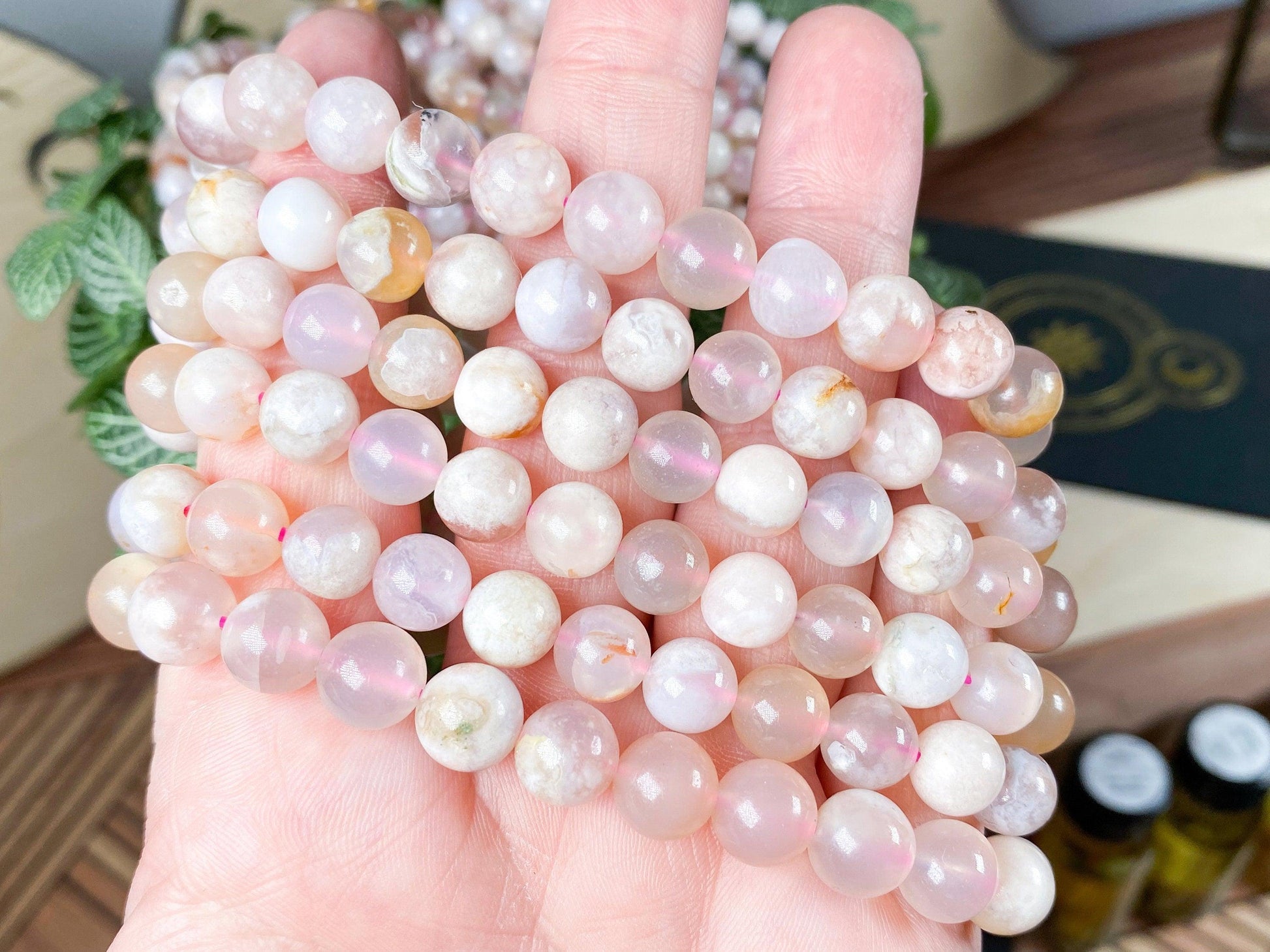 A hand holds several strands of smooth, round Cherry Blossom Flower Agate beads from The Crystalary, showcasing subtle white and yellow patterns. The beads are uniformly arranged with a glossy finish, ideal for creating Cherry Blossom Flower Agate Crystal Bracelets that resonate with the heart chakra. In the background, green foliage and a decoratively patterned item appear blurred.