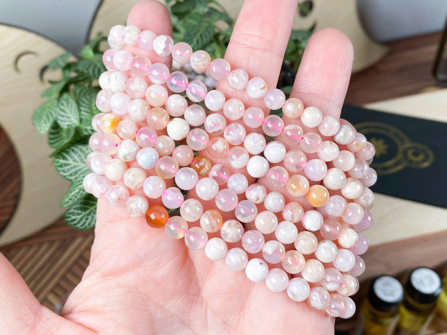 A hand holds several strands of round, semi-translucent beads in varying shades of pink and peach, resembling the Cherry Blossom Flower Agate Crystal Bracelet by The Crystalary. The background showcases a decorative surface adorned with green plants, wooden elements, small bottles, and hints at crystal bracelets designed to balance the heart chakra.