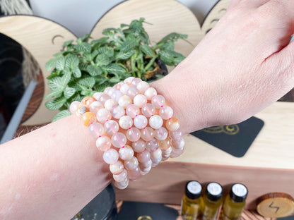 A hand is shown wearing multiple stacked beaded crystal bracelets in shades of pink and white, including The Crystalary's Cherry Blossom Flower Agate Crystal Bracelet. In the background, a wooden surface is adorned with leaf-themed decor, small bottles, and lush green plants. These bracelets are perfect for balancing the heart chakra.