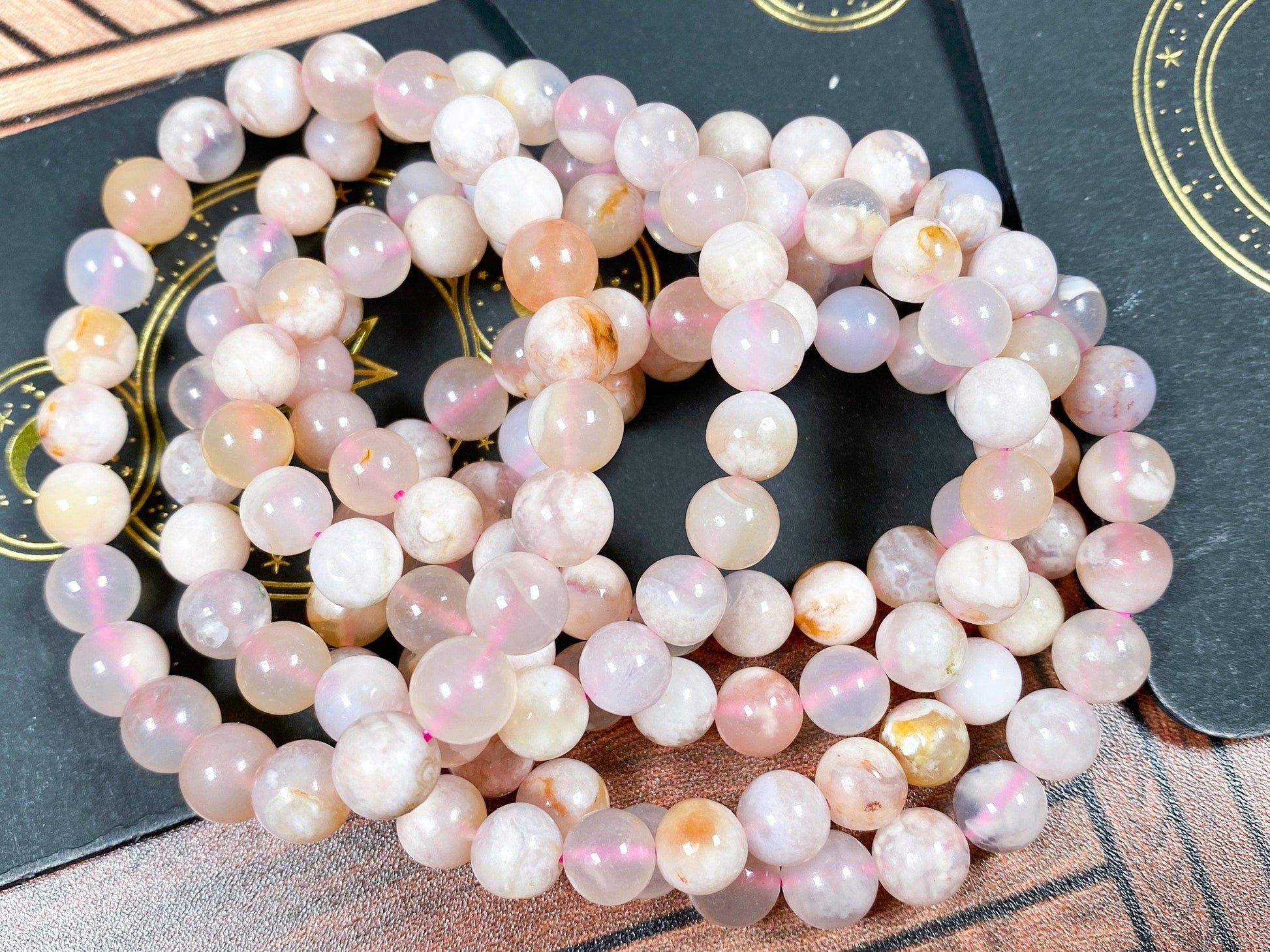 A close-up photo of several Cherry Blossom Flower Agate Crystal Bracelets by The Crystalary, made from round, polished gemstones in varying shades of pink, white, and light brown. The bracelets are arranged in loops on a wooden surface with dark cards in the background featuring gold designs, emphasizing the heart chakra energy they impart.