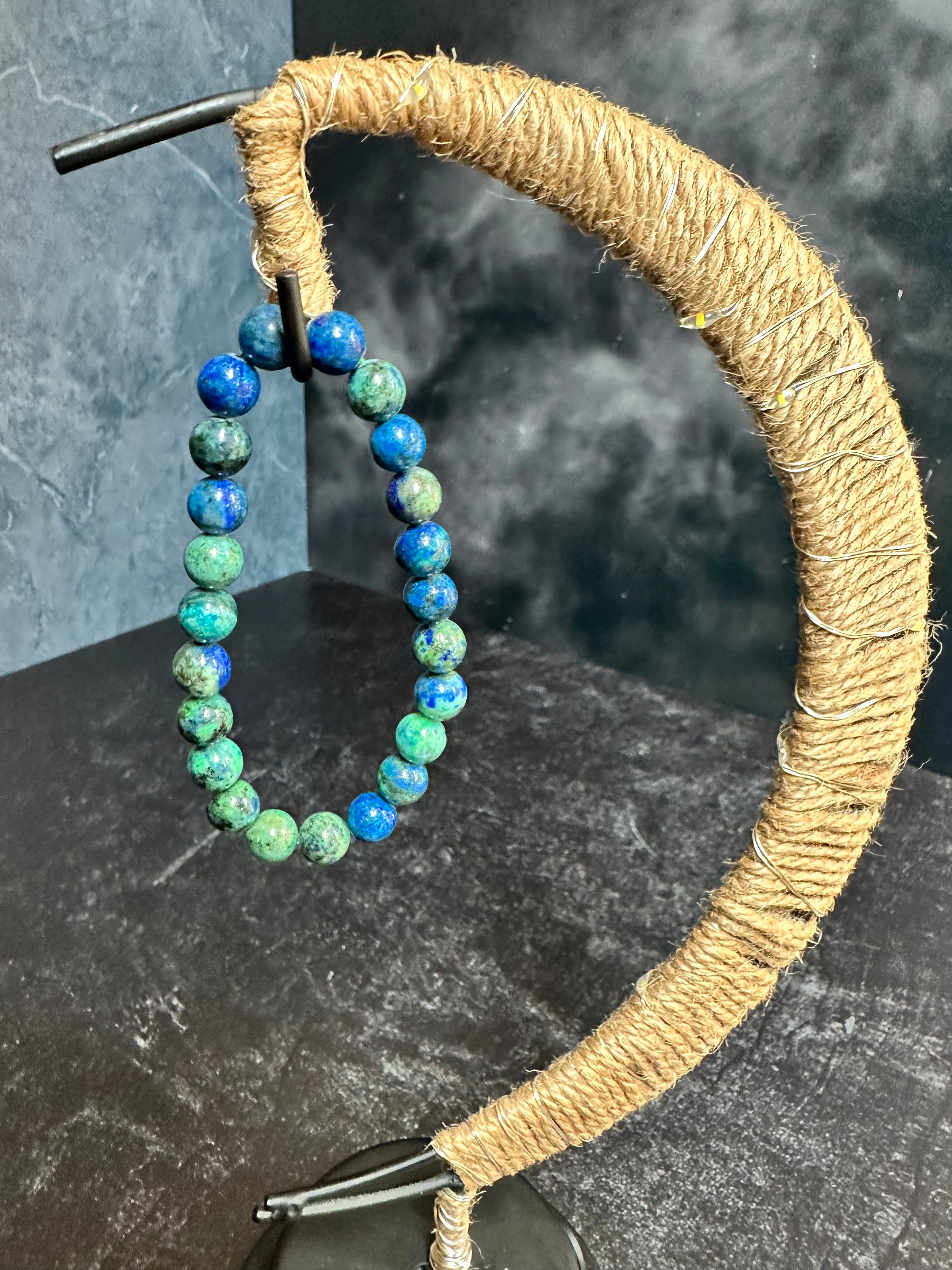 A Chrysacolla Crystal Bracelet from The Crystalary hangs on a uniquely shaped rustic display stand wrapped in twine. Crafted from ethically sourced gemstones, this intricately arched stand showcases polished stones that exude calming energy in varying shades of blue and green, set against a dark, textured background.