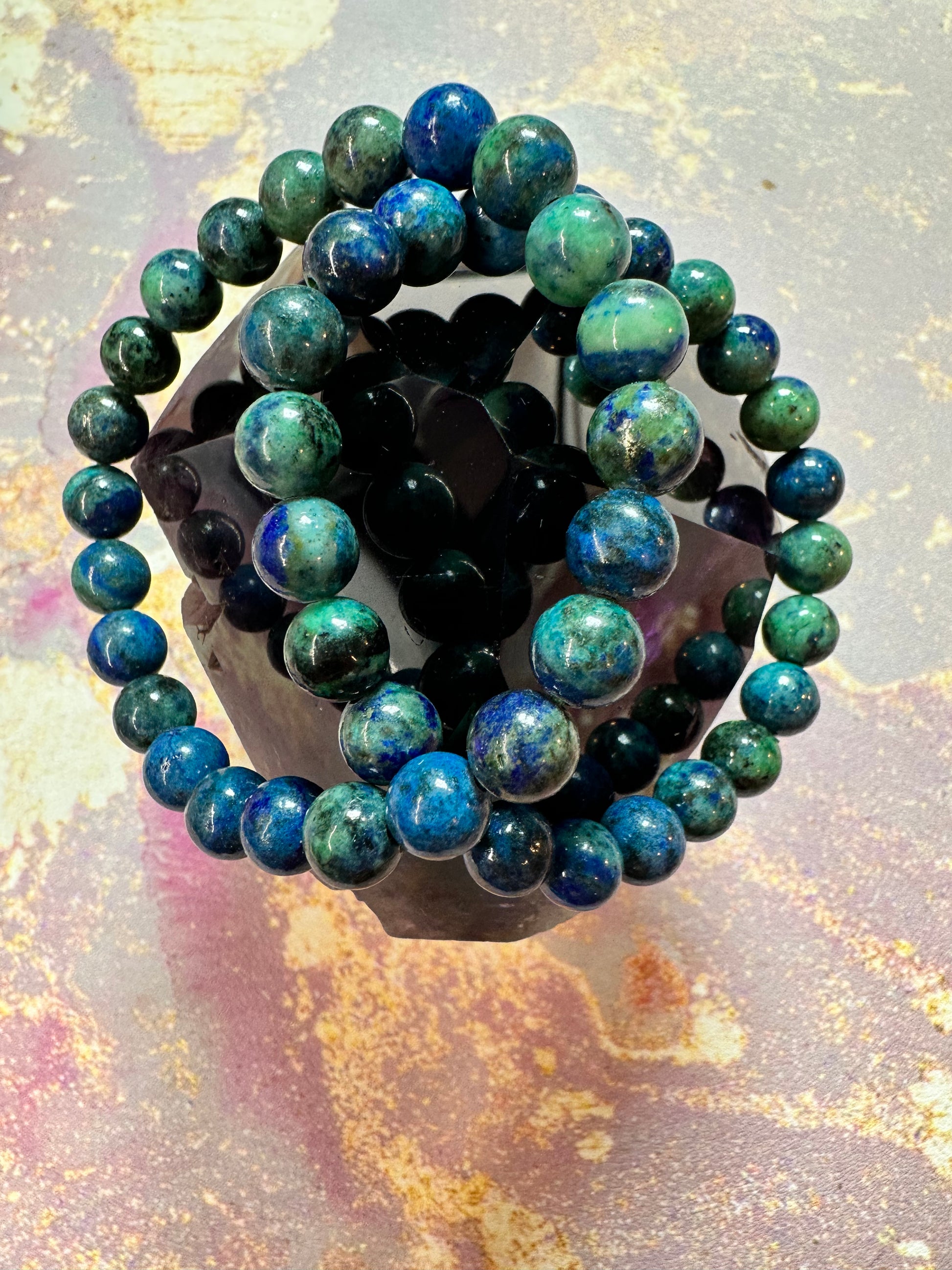 The Chrysacolla Crystal Bracelet from The Crystalary, featuring round, blue and green marbled beads, is elegantly displayed on a dark, flower-shaped crystal stand. The backdrop showcases a vibrant abstract design with splashes of pink, orange, and purple hues. This exquisite piece emanates calming energy and promotes the ethical crystal trade.