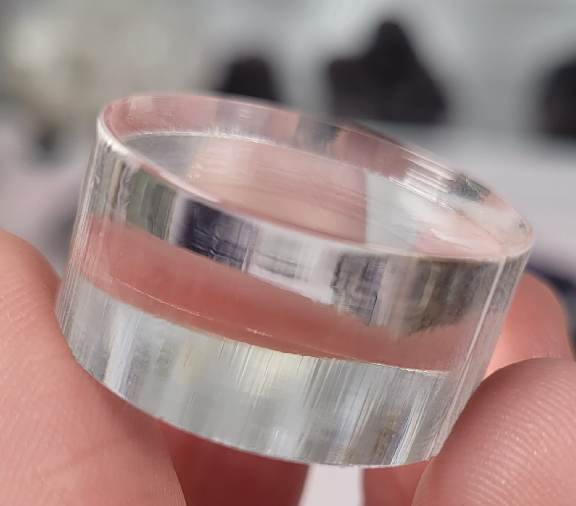 Close-up image of a person holding a transparent cylindrical object using their thumb and finger. The object has clear, smooth surfaces that reflect and refract the surrounding light, creating a distortion of the background, similar to how items are showcased on The Crystalary's Circle Acrylic Stand/Mount 1" x 1/2".