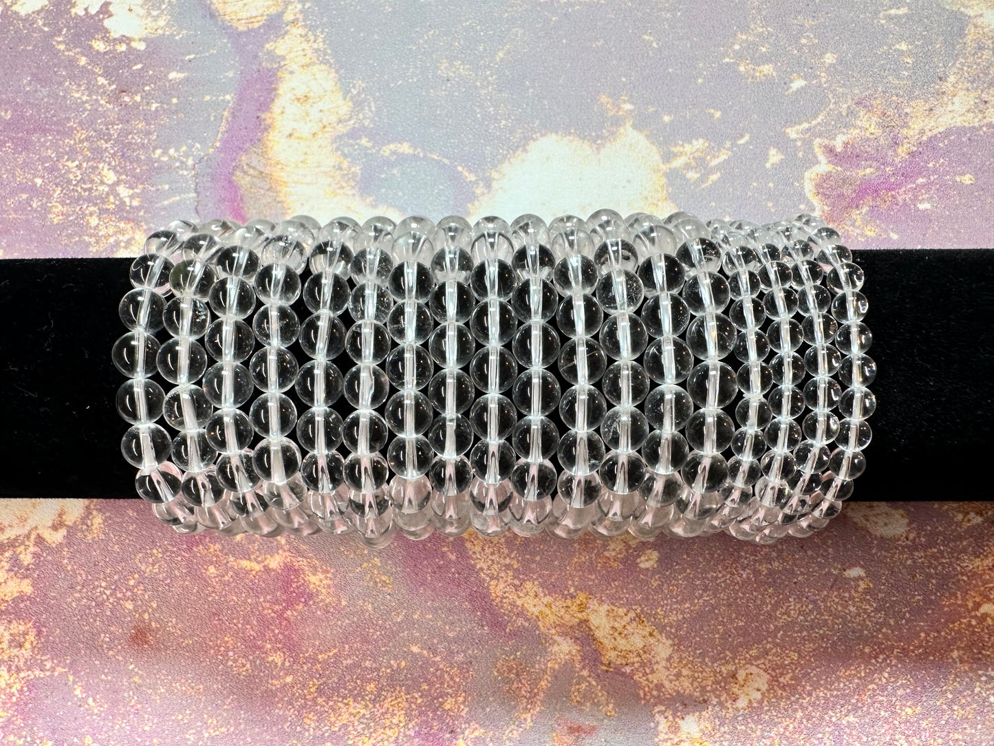 A series of clear quartz crystal beads from The Crystalary are strung together and placed on a black rod, forming multiple intertwined loops reminiscent of elegant Clear Quartz Crystal Bracelets. The background has a marbled texture in soft pink, purple, and yellow hues, adding a subtle contrast to the clear beads.