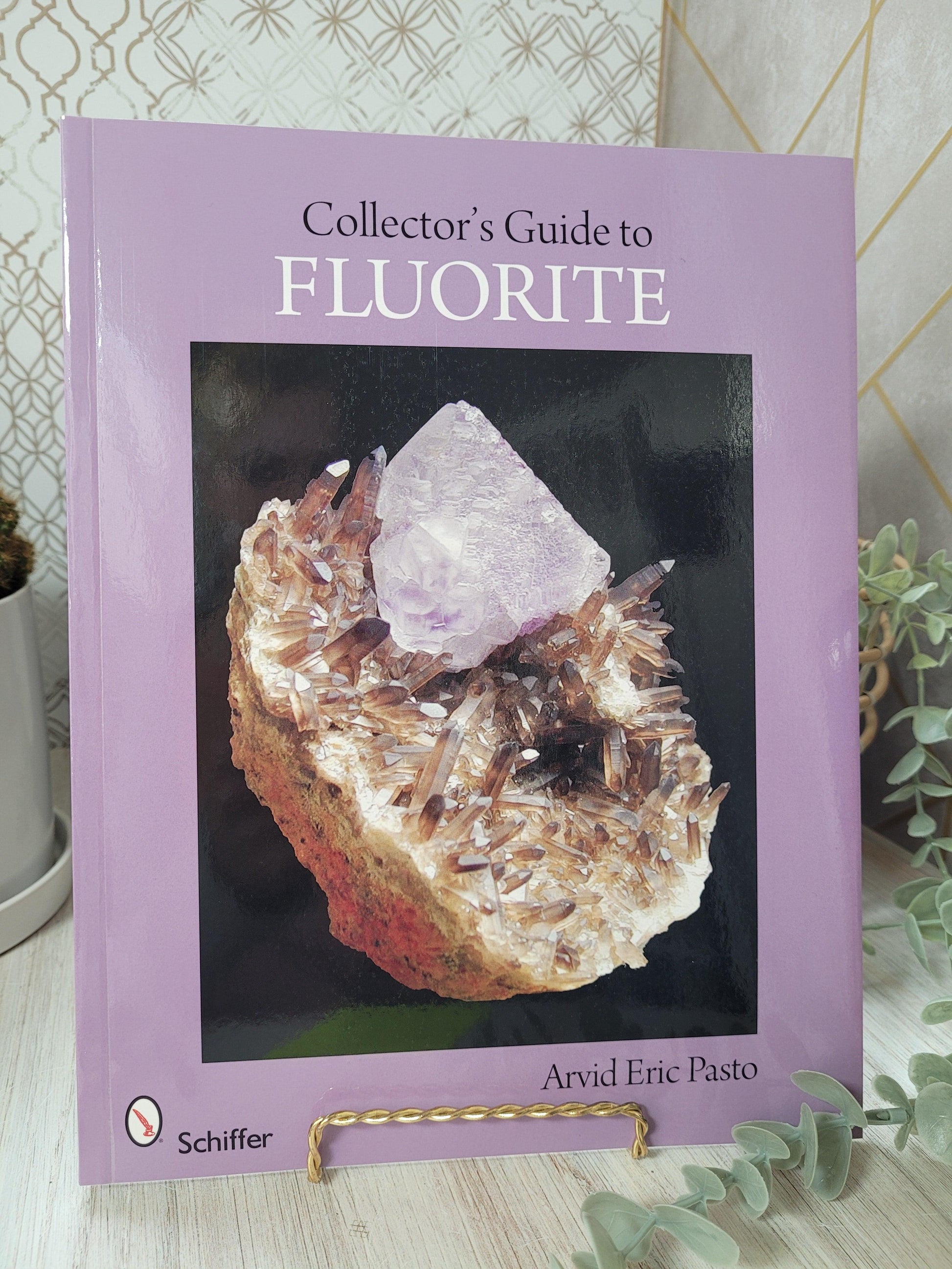 A book titled "Collector's Guide to Fluorite by Arvid Eric Pasto" from The Crystalary features a light purple cover with a prominent fluorite crystal on a matrix of smaller crystals. This indispensable guide for any rockhound is displayed on a stand with a plant in the background.