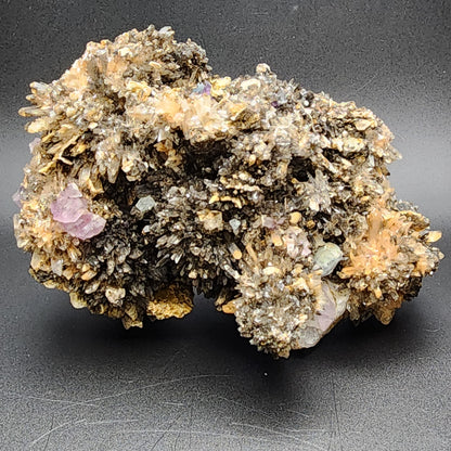 A close-up of a complex, multi-hued Creedite cluster from Navidad Mine in Inde Municipality, Durango, Mexico against a dark background. The specimen from The Crystalary features numerous sharply pointed crystals ranging in color from golden-brown to purple, creating an intricate and textured appearance with high fluorescence.