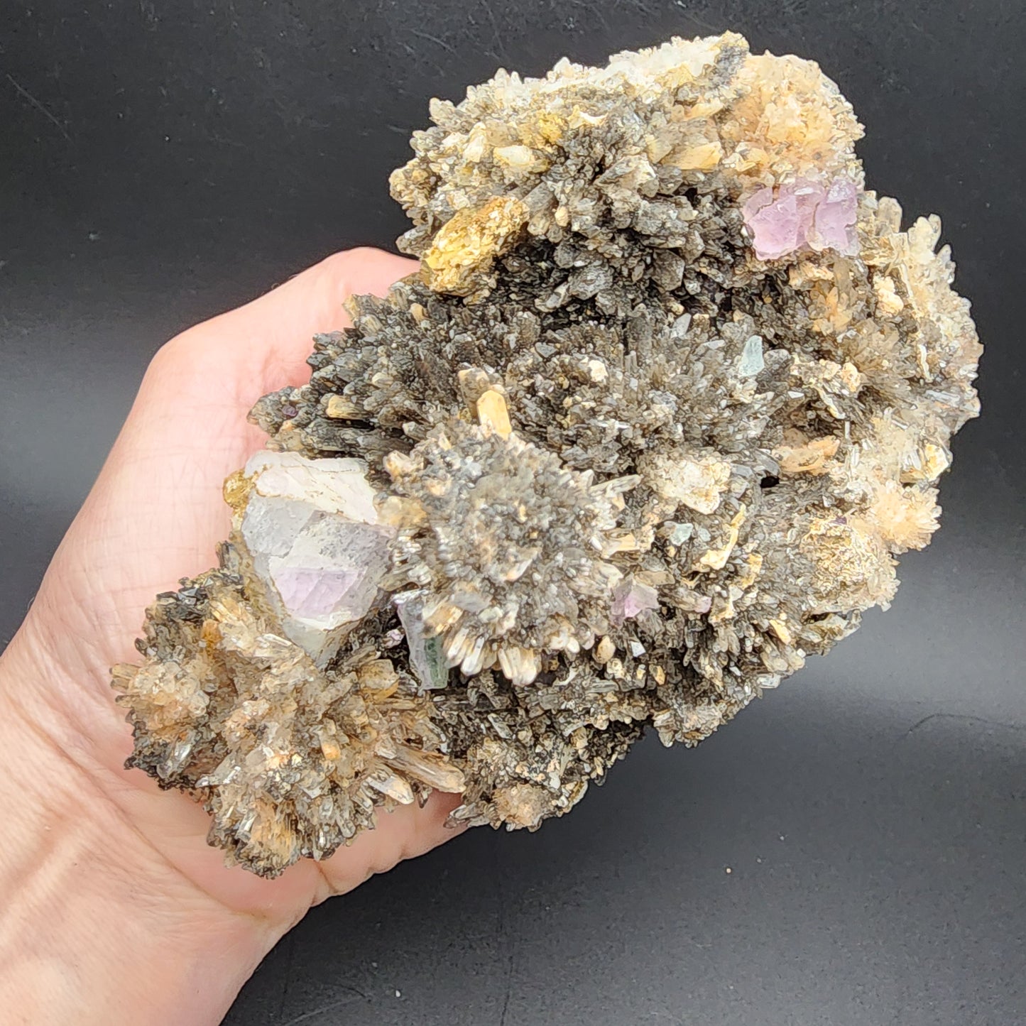 A hand holds a large, rugged Creedite and Fluorite cluster from Navidad Mine in Inde Municipality, Durango, Mexico, featuring numerous spiky, grayish-brown formations with small patches of light purple and clear crystallized fluorite areas. The solid black background of the image sharply contrasts with the textured and colorful mineral specimen from The Crystalary.