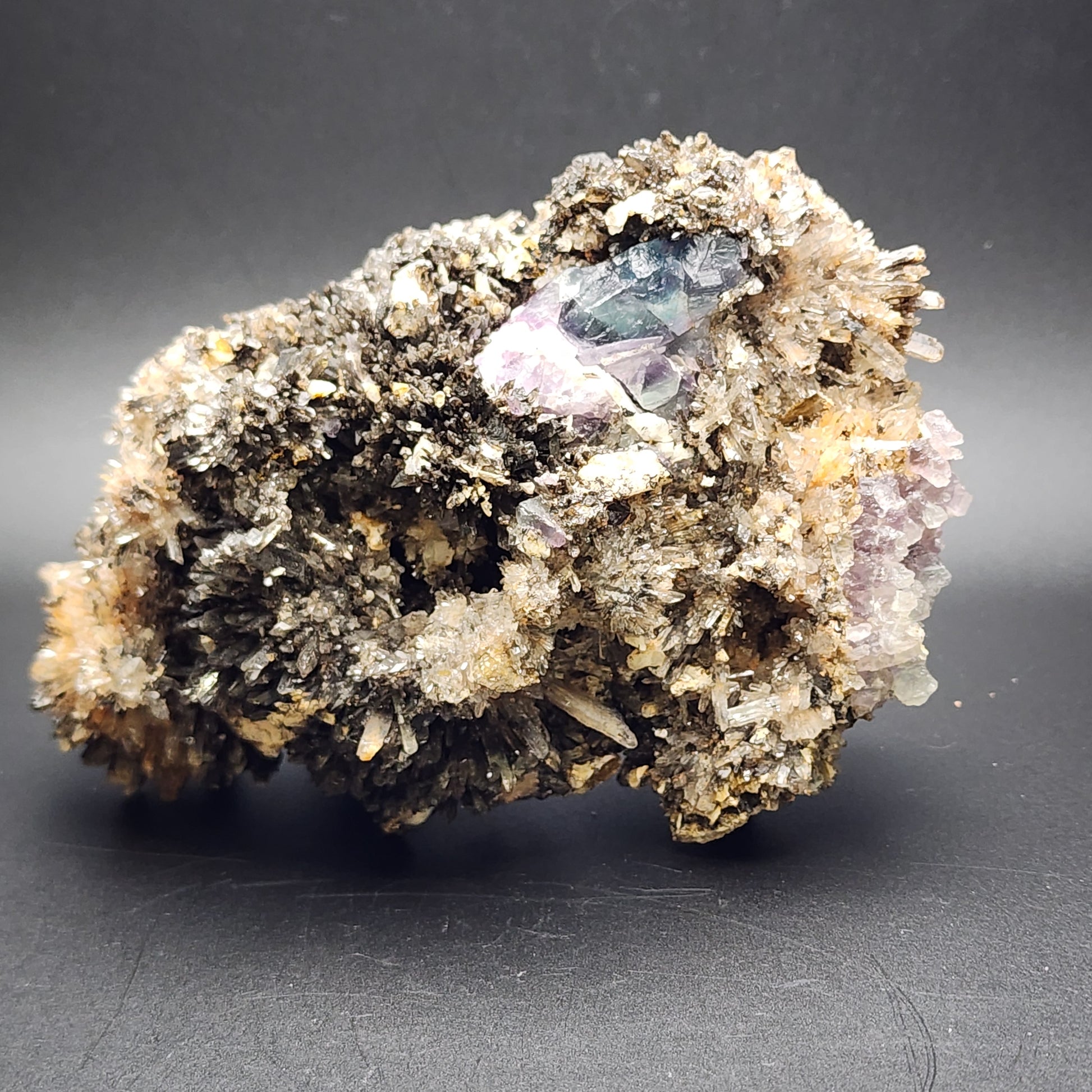 A large, intricate Creedite mineral rock from the Navidad Mine in Inde Municipality, Durango, Mexico, displays a rough surface with clusters of dark crystalline formations interspersed with patches of lighter translucent crystals. Notably featured is a section of purple Fluorite embedded within. The backdrop is a smooth, dark surface. This stunning piece is brought to you by The Crystalary.