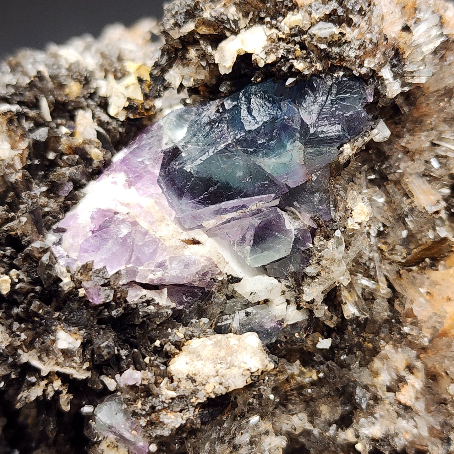 Close-up image of a mineral formation showcasing a cluster of multicolored Creedite crystals from the Navidad Mine in the Inde Municipality, Durango, Mexico. The crystals, primarily purple and blue Fluorite, are surrounded by textured and fragmented materials, including grains and smaller stones. The intricate layers and high fluorescence create a striking geological pattern. Available from The Crystalary.
