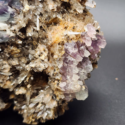 Close-up of a mineral specimen from The Crystalary, featuring clusters of translucent purple fluorite and clear crystals against a dark background. This piece, known as Creedite and Fluorite from the Navidad Mine in Inde Municipality, Durango, Mexico, showcases high fluorescence crystals with a jagged, spiky appearance surrounded by earthy brown and grey tones.