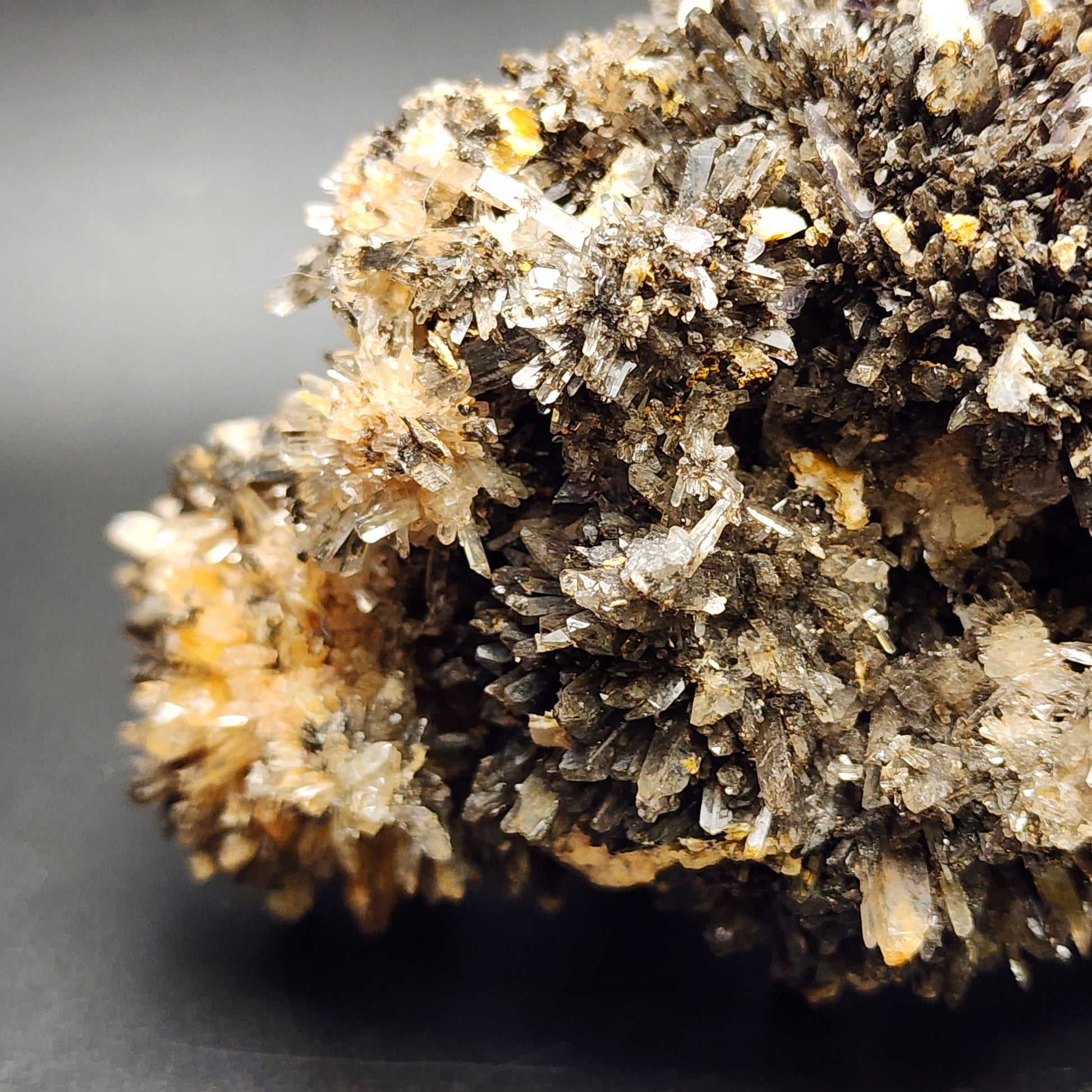 A close-up image showcases a cluster of spiky orange creedite formations, featuring dark gray, black, and tan hues. The detailed crystal structures exhibit an array of small, sharp points against a solid, dark background. This beautiful specimen originates from the Navidad Mine in Inde Municipality, Durango, Mexico and is offered by The Crystalary under the name Creedite with Fluorite.