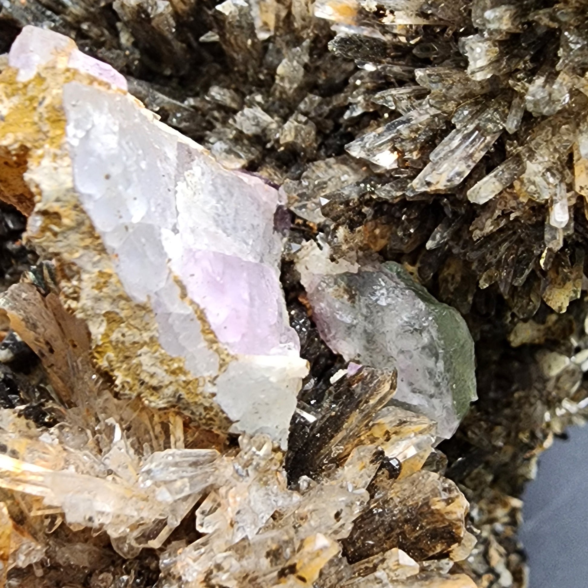 A close-up of a crystalline mineral formation featuring clusters of clear, spiky quartz crystals surrounding chunks of raw fluorite in captivating shades of purple, green, and white. Among them, striking orange creedite from the Navidad Mine in Inde Municipality, Durango, Mexico adds an intriguing contrast. The textures are rough and jagged, with intricate details on the surface. This exquisite piece is brought to you by The Crystalary.