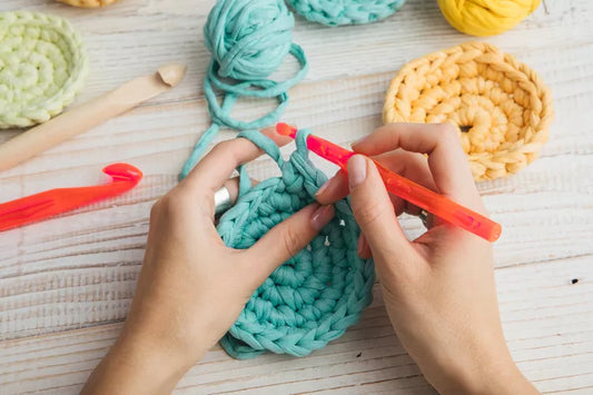 Hands crochet a blue circle with chunky yarn, encircled by colorful yarn balls and a crochet hook on wood. This vibrant project is akin to the creative artistry at The Crystalary’s Crochet Class on May 3, 2 PM EST in Cincinnati.