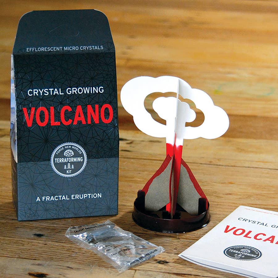 A "Crystal Growing Volcano" craft kit from Copernicus Toys sits on a wooden surface. Perfect as science gifts, it includes a volcano model with a red and white design, an instruction booklet, a sachet of chemicals, and a decorative cardboard backdrop titled "Crystal Growing Volcano: A Fractal Eruption.